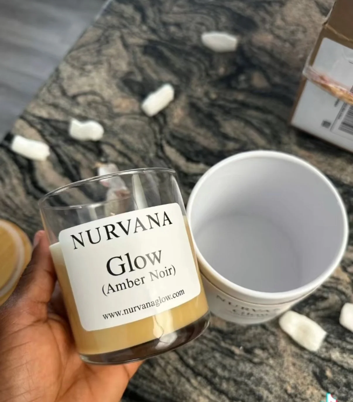 A hand holding a lit candle labeled 'Nurvana Glow Amber Noir' with an empty white container and scattered white wax melts on a dark granite countertop.