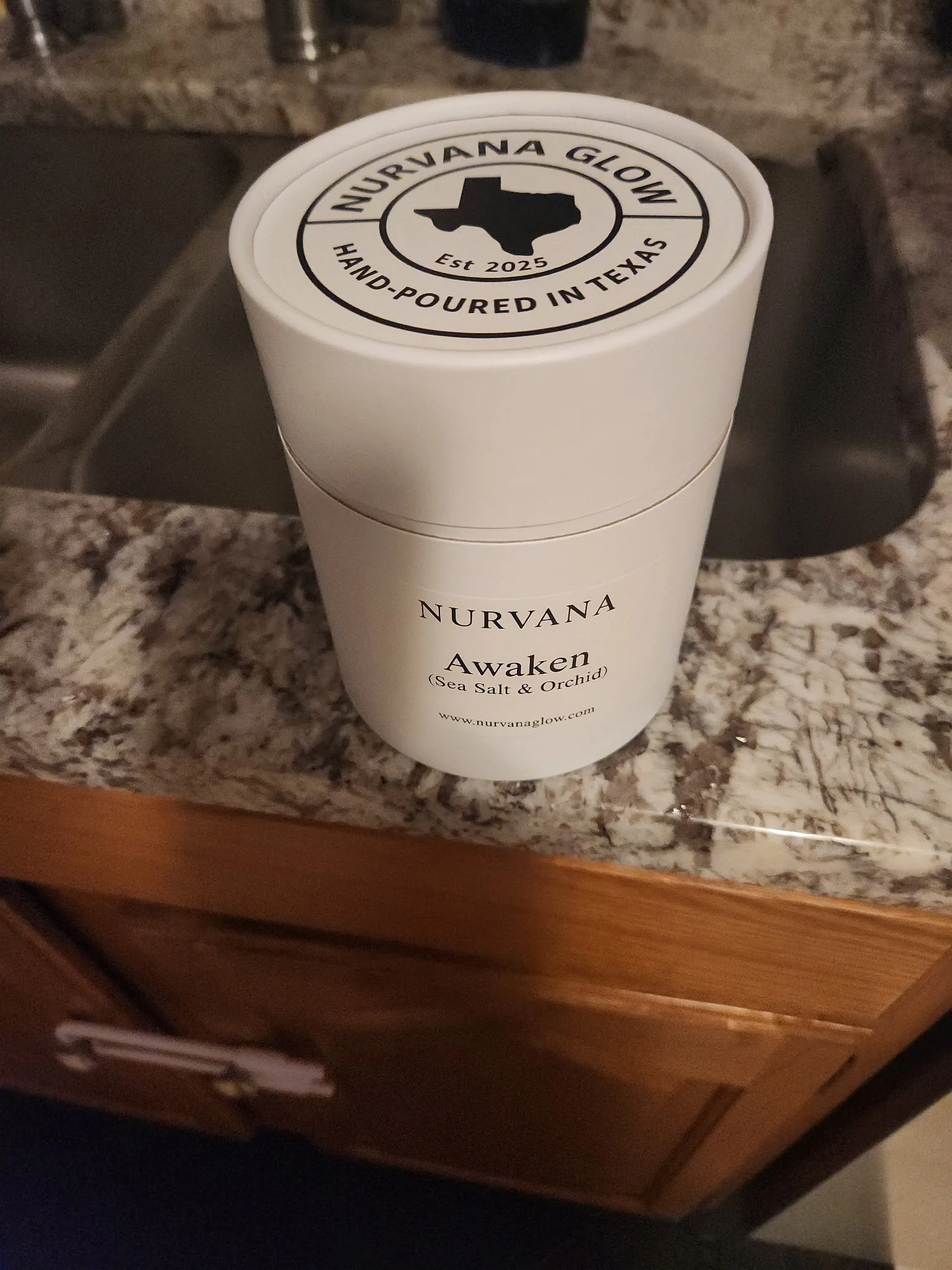 A white container with a black and white label, sitting on a granite countertop. The label reads "NURVANA Awaken (Sea Salt & Orchid)" and the top of the container has a logo with a Texas-shaped outline, the text "NURVANA GLOW," and "Est 2025."