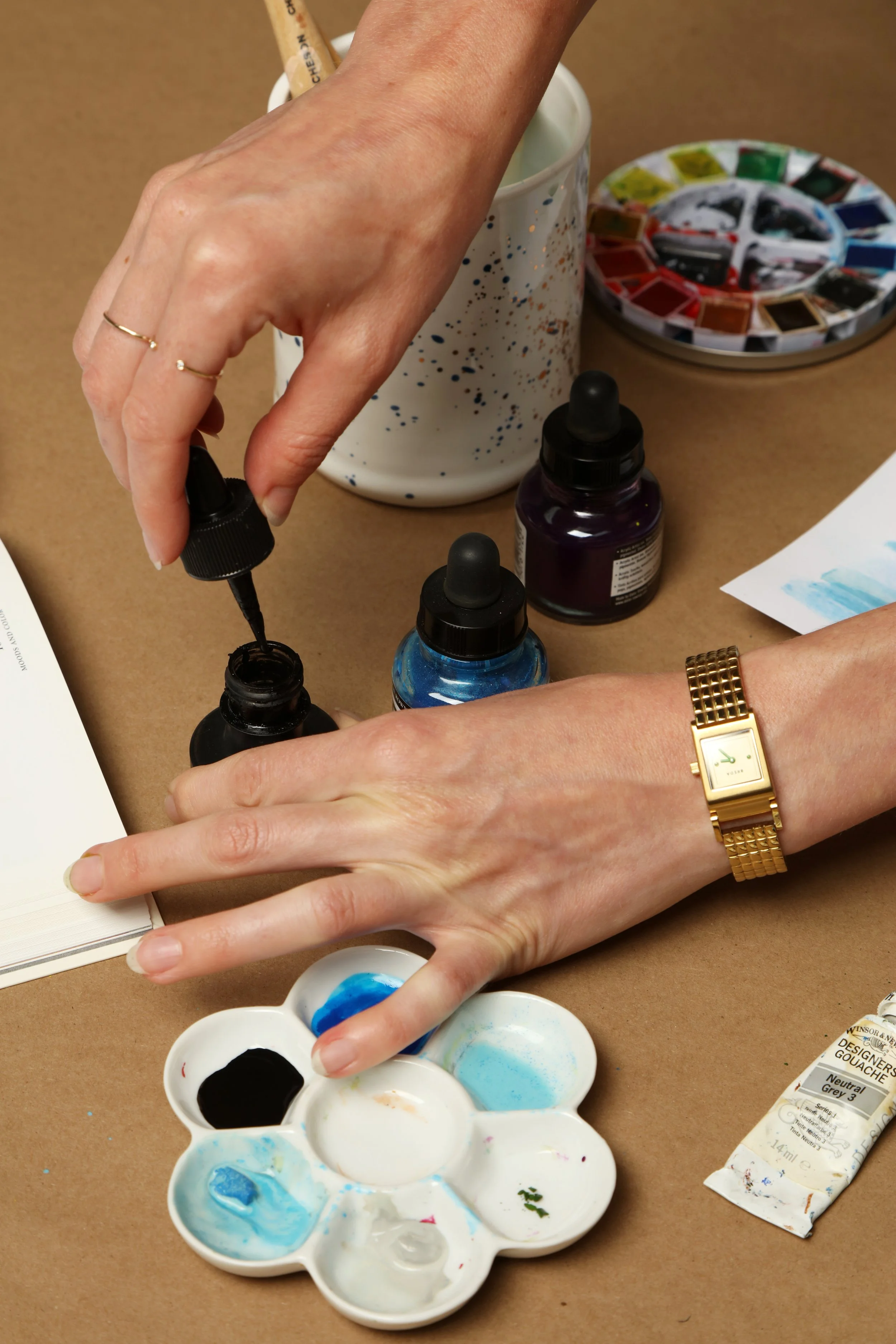 Artist preparing black ink with a dropper surrounded by watercolor tools, palette, and paint jars on a craft table.