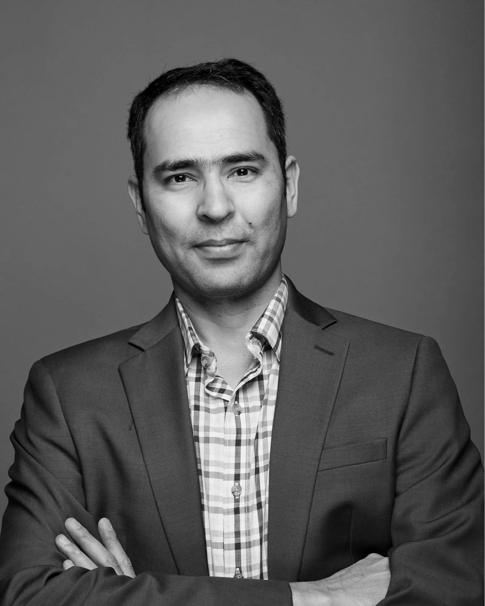 Black and white portrait of a man in a suit with crossed arms, wearing a checkered shirt, looking directly at the camera with a neutral expression.
