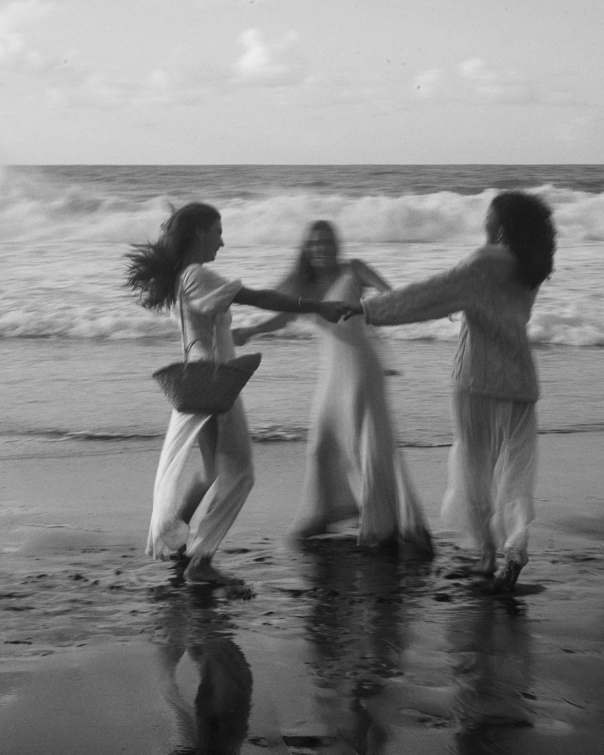 Three people on the beach holding hands, with ocean waves in the background, black and white photograph.