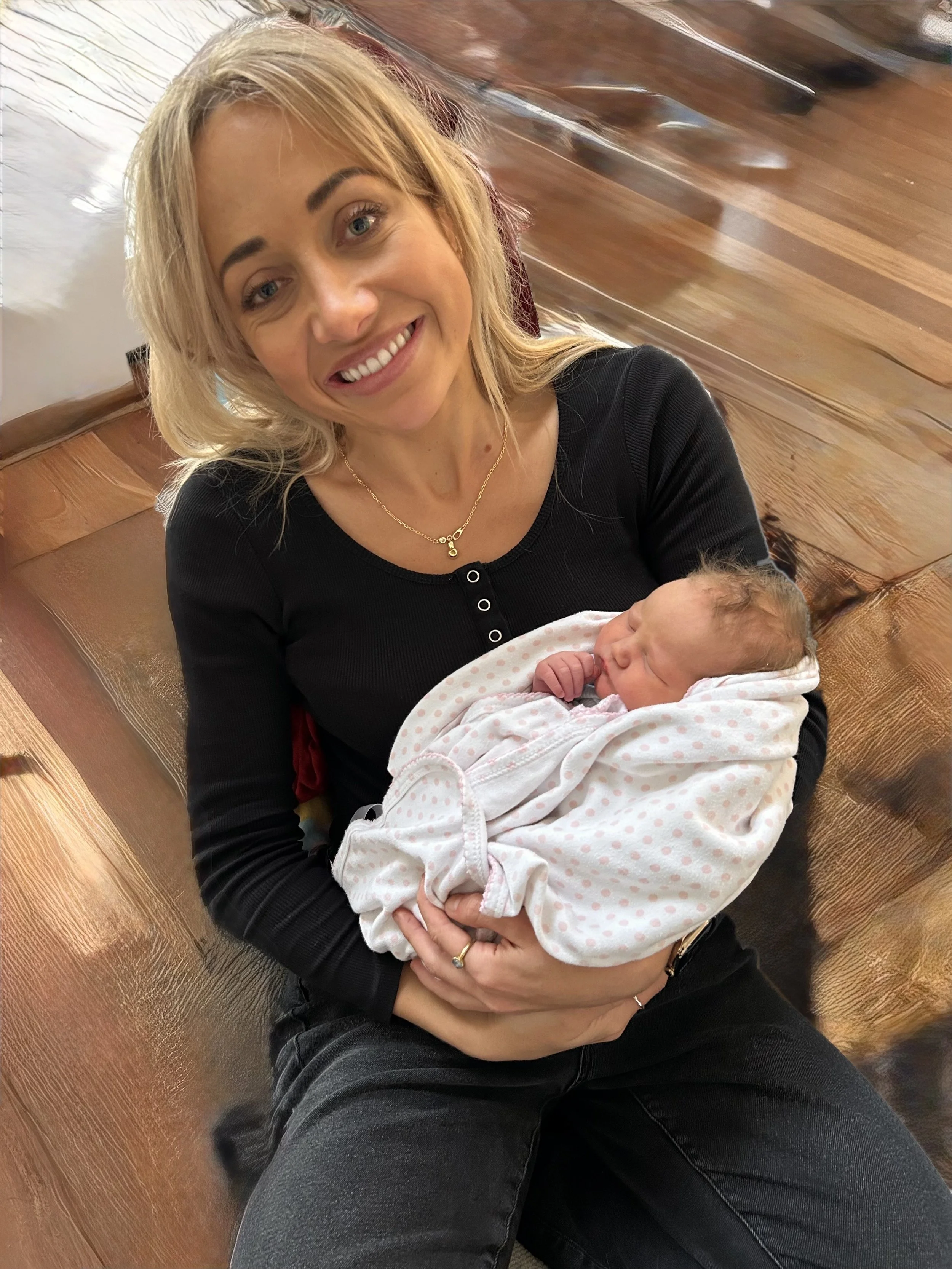 A woman with blonde hair smiling while holding a newborn wrapped in a white blanket with pink polka dots, seated on a wooden floor.