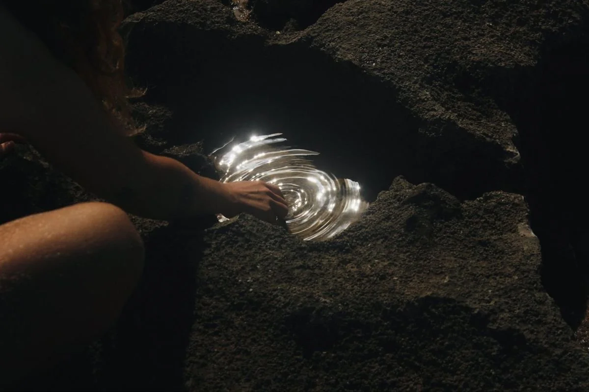 A person touching water in a dark, rocky cave, creating ripples as the water reflects light.