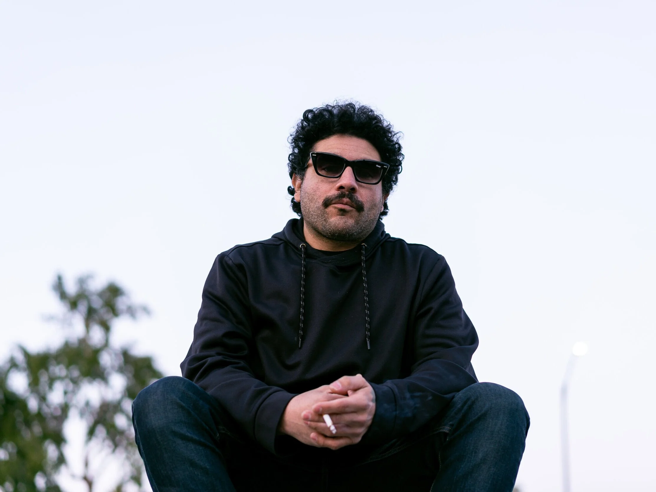 Artist Philthy Dronez is squatting, sporting sunglasses on his face and a cigarette in his folded hands.