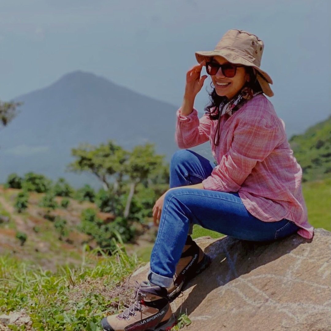 Stephanie Segovia is sitting and smiling on a rock outdoors. Mountains are seen in the background, hinting at her growing up "surrounded by coffee mountains."