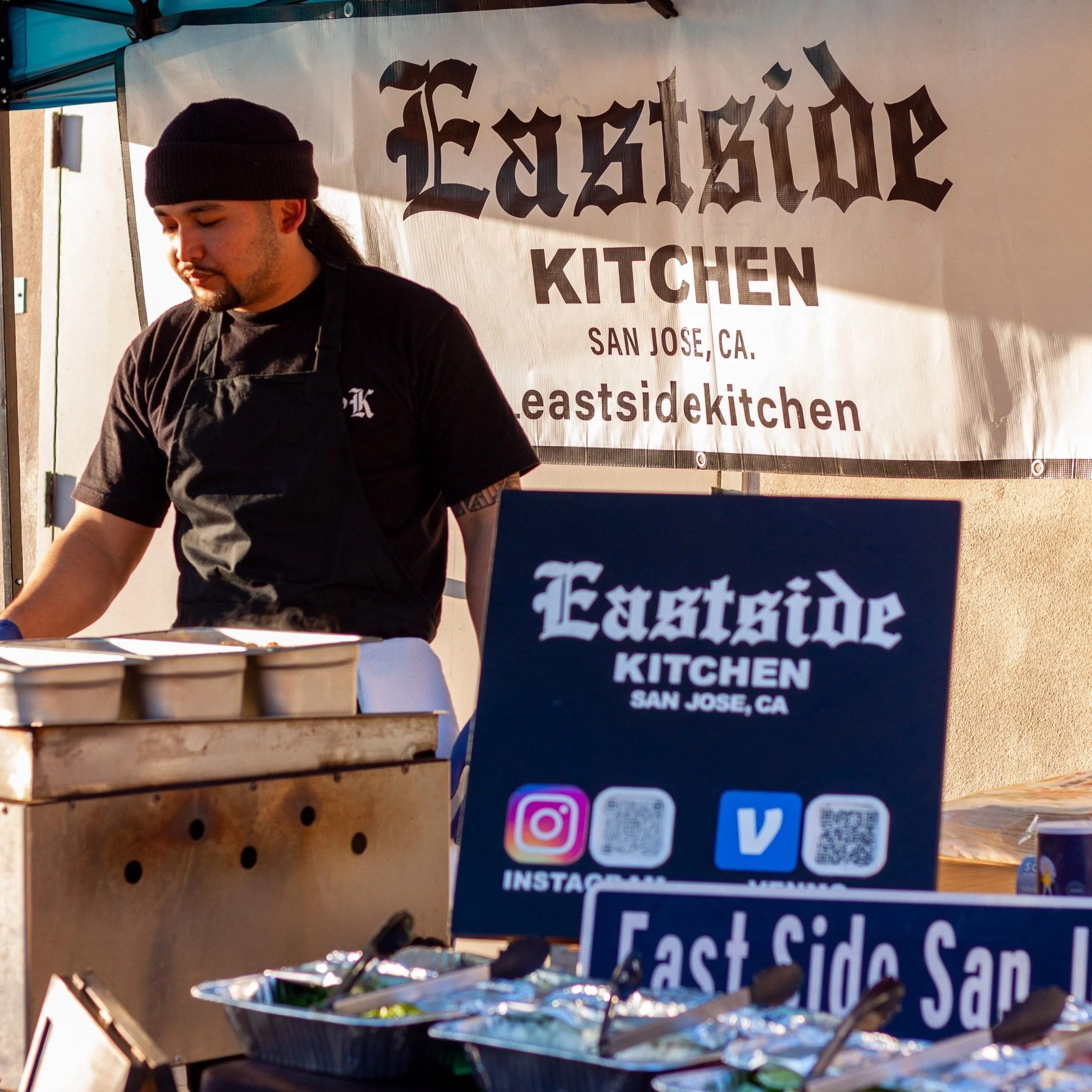 Jojoe Delgado is pictured in a beanie and apron and is looking down at an array of trays of food in front of him. Two signs read "Eastside Kitchen, San Jose, CA."