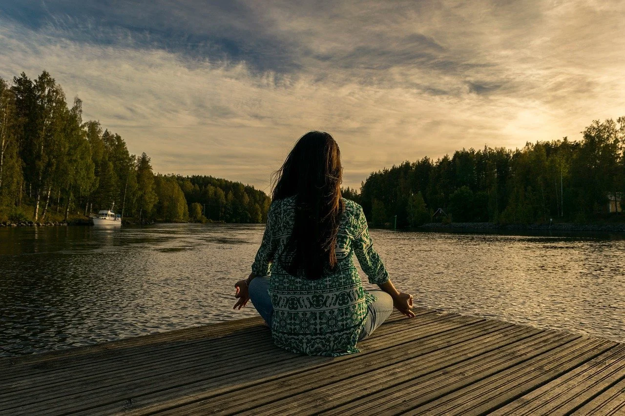 A woman with long dark hair sitting cross-legged on a wooden dock by a river, facing the water with trees on both sides, during sunset.