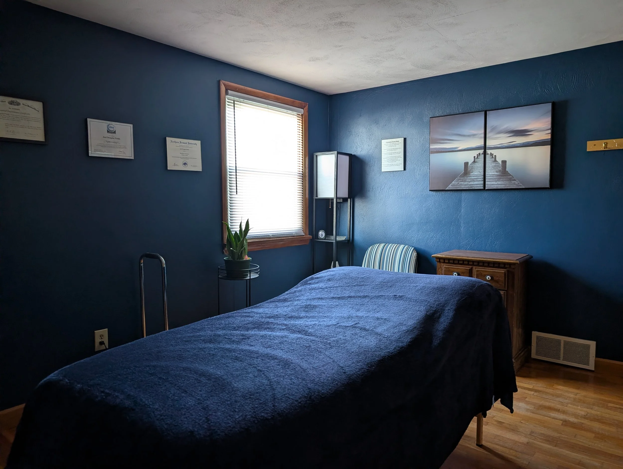 A bedroom with a blue wall, a window with blinds, a bed with a dark blue cover, a wooden nightstand, a striped chair, a plant on a stand, and framed certificates on the wall.