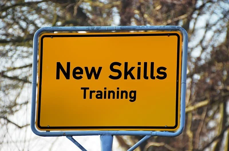 A sign that reads "New Skills Training" in black text on a yellow background, with trees in the blurry background.