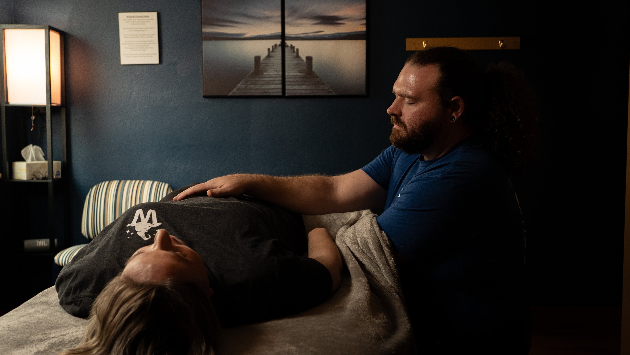 A man with a beard and long dark hair, wearing a blue shirt, is performing acupuncture on a person lying face up on a treatment table. The acupuncture includes thin needles in the person's arm and chest. The setting appears to be a professional clinic with a dark wall, a framed photo of a pier leading into a body of water, and a shelf with various items.