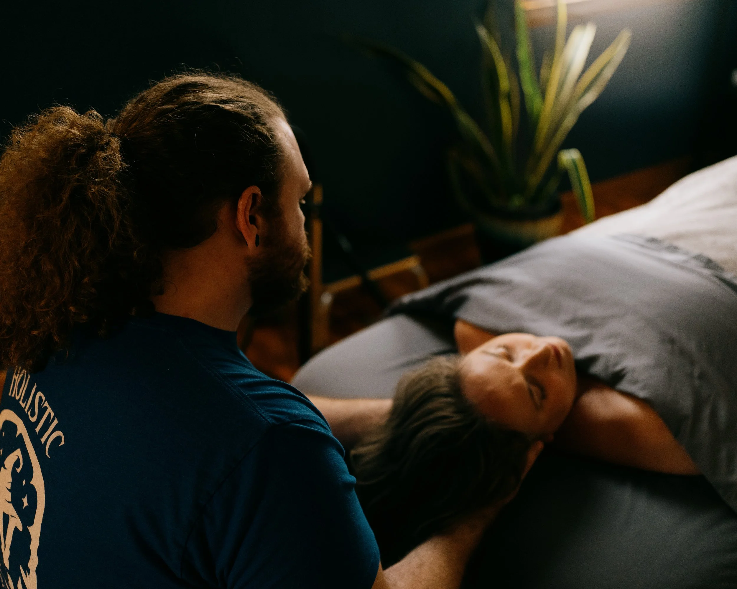 A person with long, curly hair and earrings, wearing a dark T-shirt, is comforting a woman lying on a bed. The woman is resting her head on her arm, with closed eyes, and is covered with a gray blanket. There is a green potted plant in the background near a dark wall.