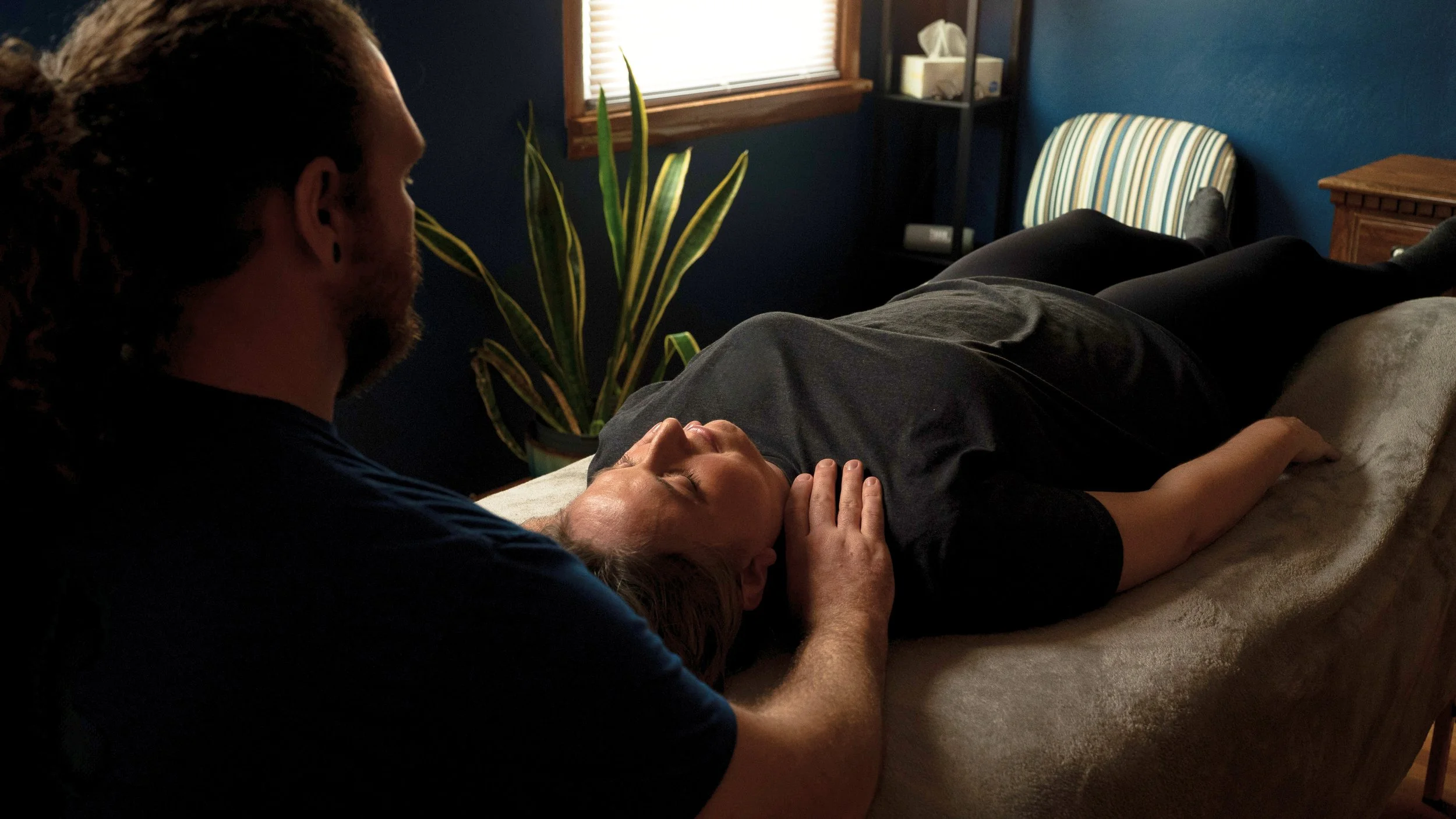 A man giving a gentle massage or relaxation therapy to a woman lying on a massage table in a cozy room with a blue wall, window, potted plant, and striped chair.