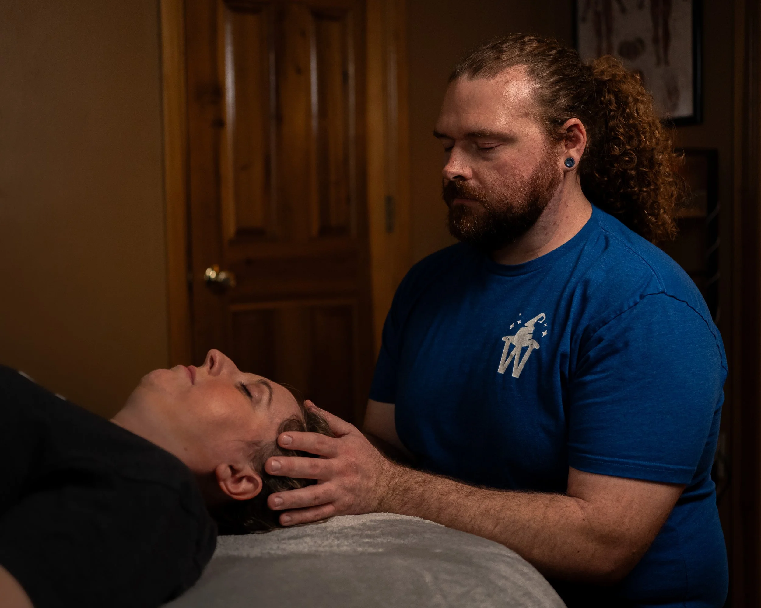 A man with curly hair and a beard is performing a chiropractic or physical therapy adjustment on another person's neck as they lie on a table with their eyes closed.