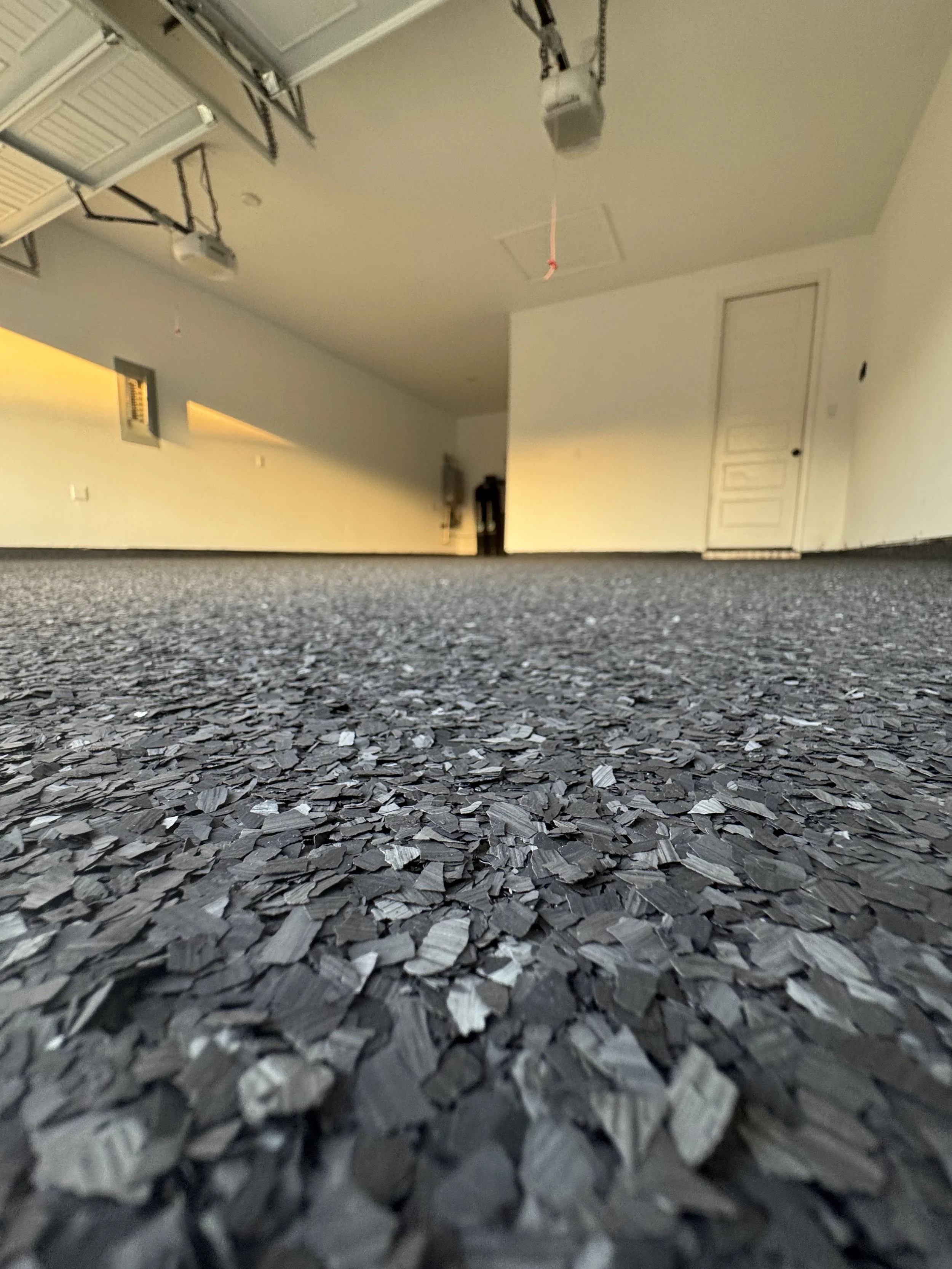 View of a garage with a textured black and gray epoxy-coated floor, white walls, a closed white door, and garage doors open on the ceiling. The garage is empty, and the photo is taken from a low angle, close to the ground.