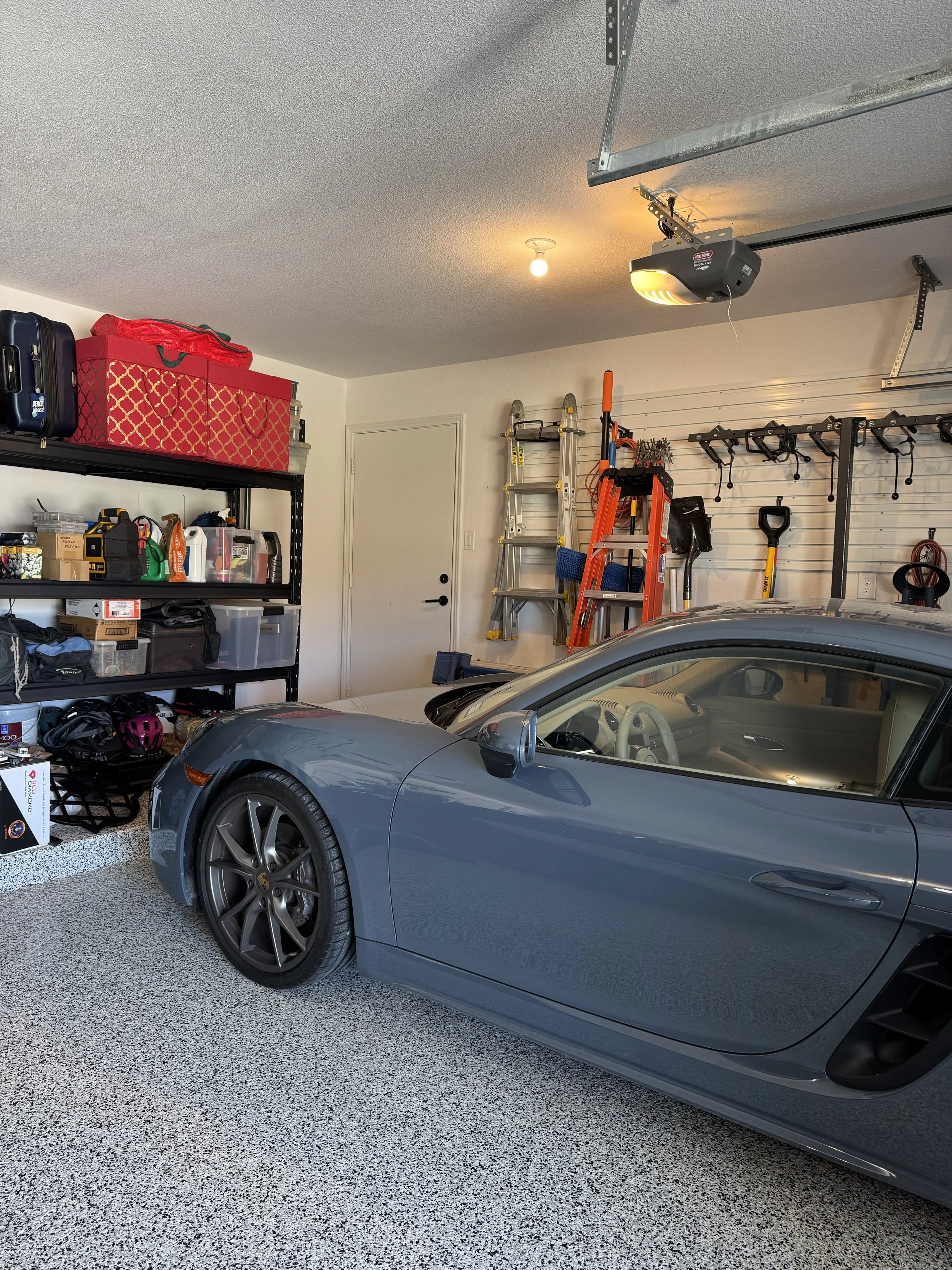 A garage with a gray sports car parked inside, containing shelves with tools, storage bins, a ladder, a garage door opener, and various gardening and maintenance equipment, with a closed white door in the background.