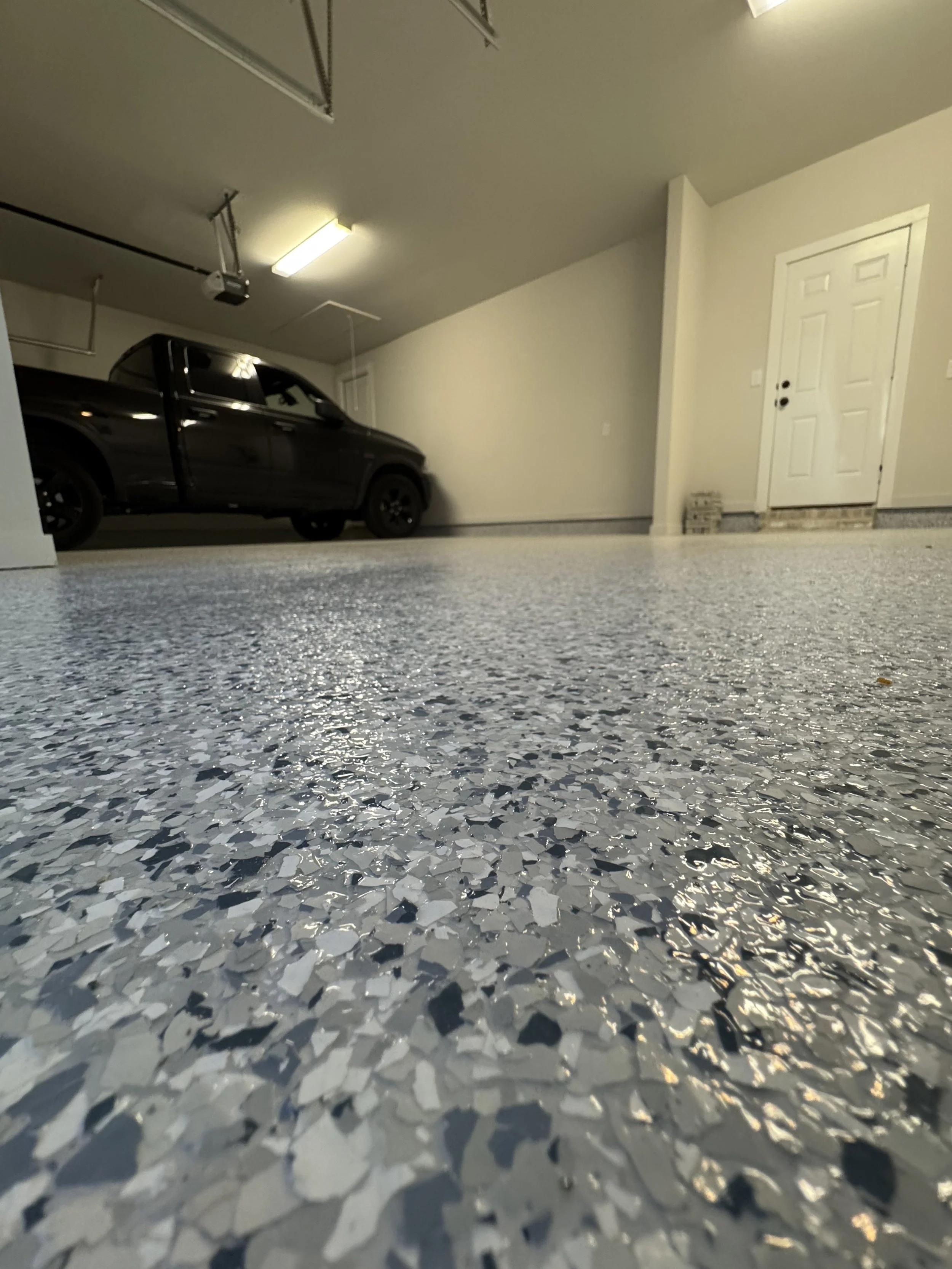 Indoor garage with a black pickup truck parked near a white door, terrazzo flooring, overhead lighting, and garage storage rails.