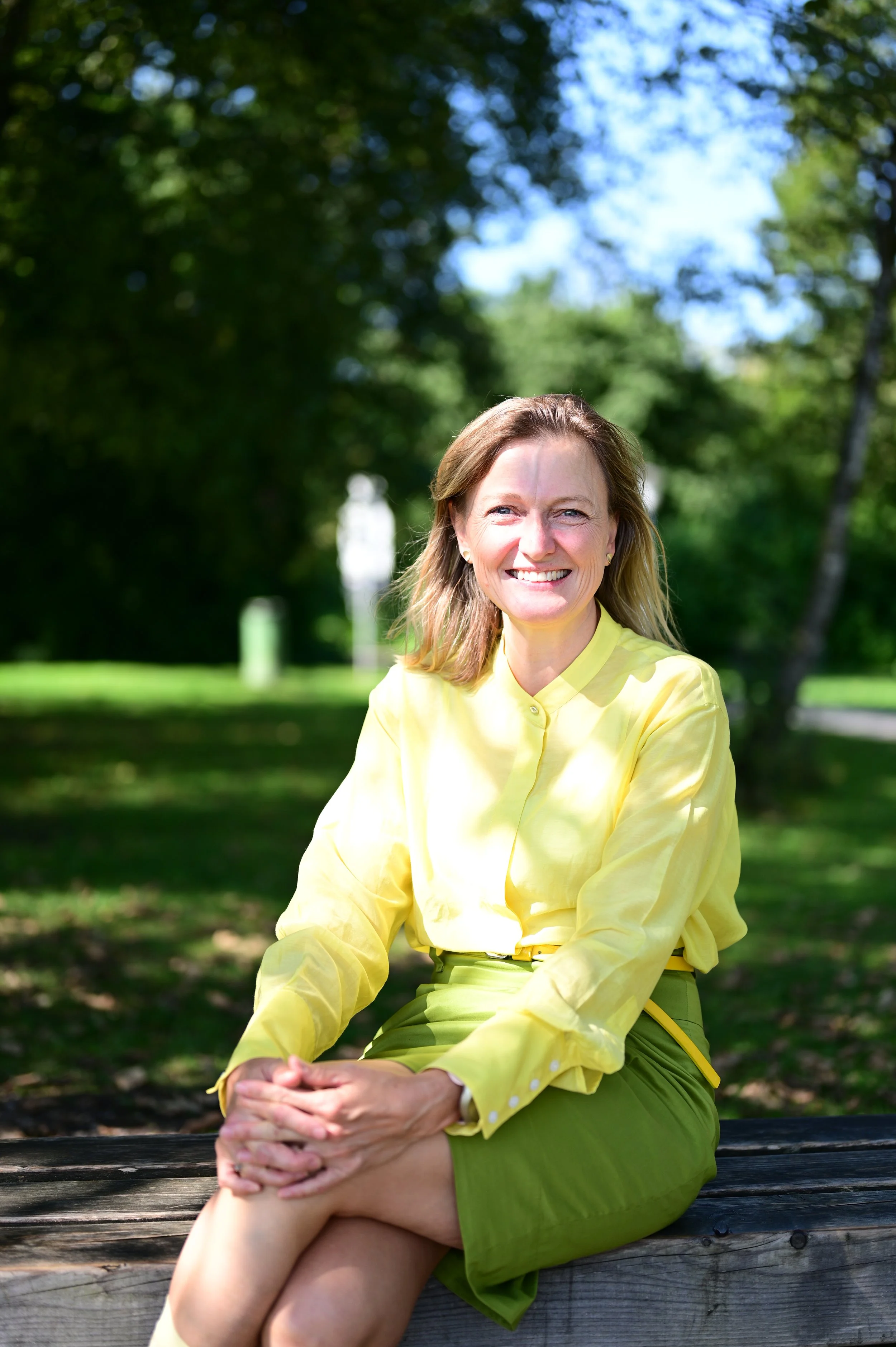 Lächelnde Frau in gelber Bluse und grüner Rock sitzt auf einer Holzbank im Park an einem sonnigen Tag.