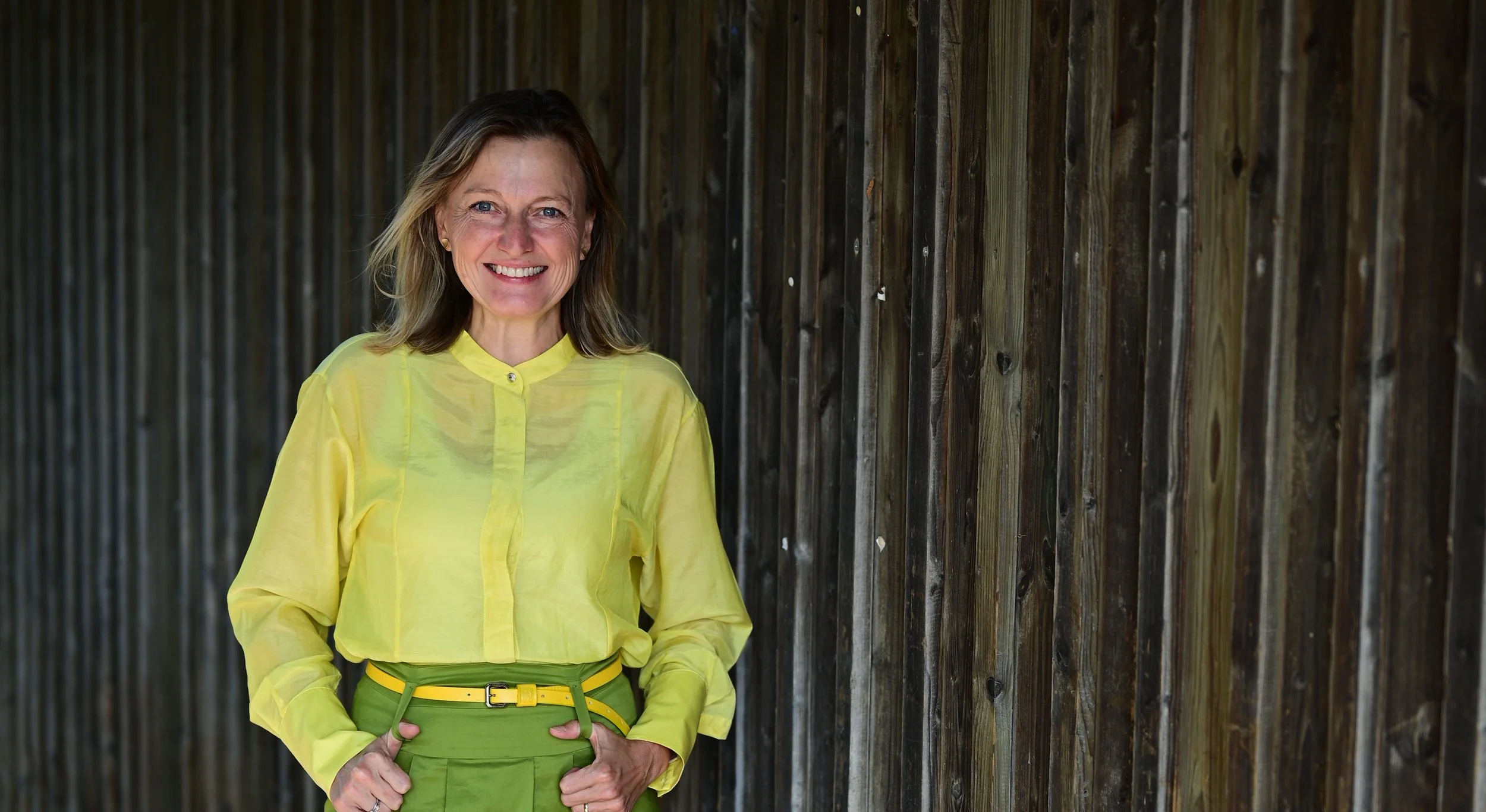 Eine lächelnde Frau in einer gelben Bluse und grünen Hosen steht vor einer Holzlattenwand.