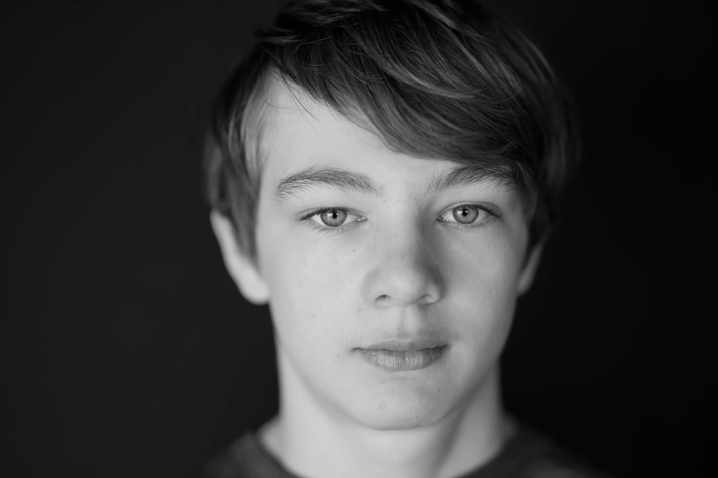 School photo of a highschooler taken in black and white near wasilla alaska. Boy is wearing a grey t-shirt with very beautiful eyes. He is wearing a serious expression.