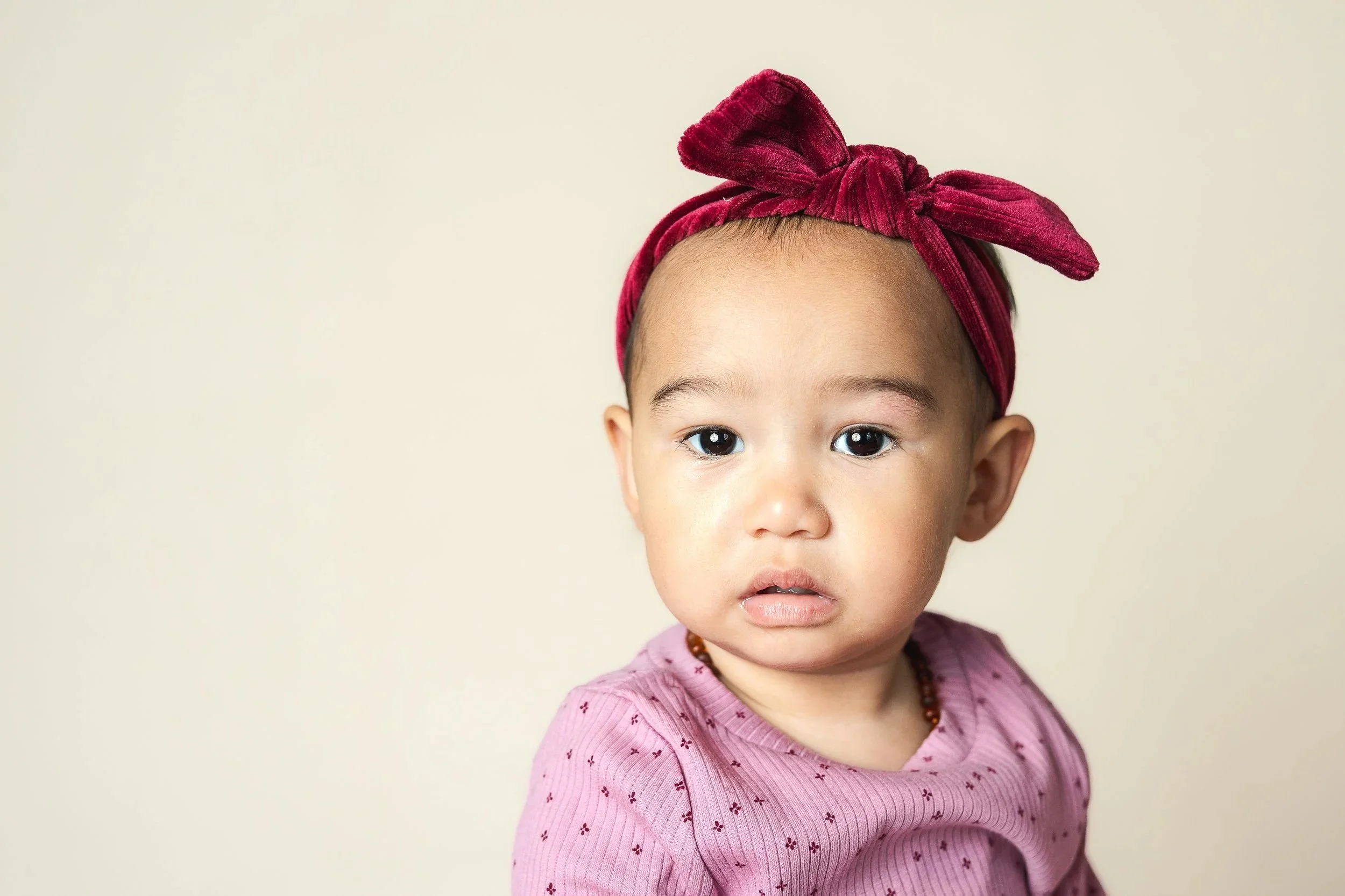Image of a baby taken at a daycare school portrait session in Anchorage Alaska. Baby 10 months old of asian descent wearing a rd bow and pink shirt on tan background.