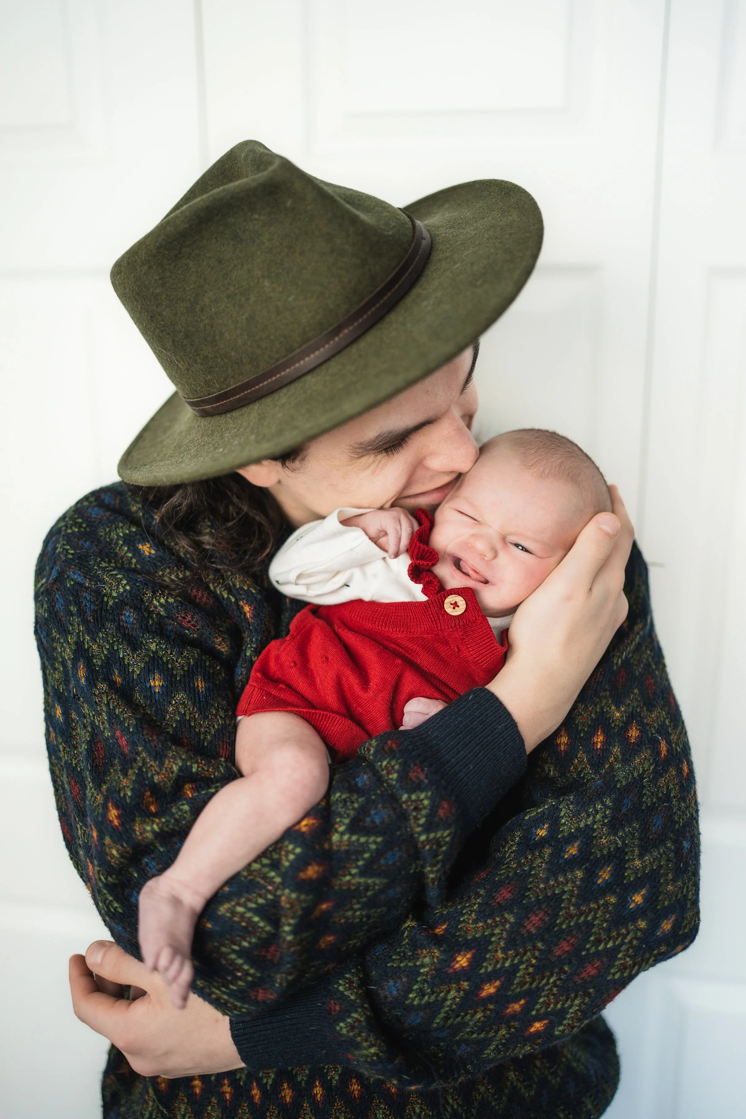 A person wearing a green hat and a colorful sweater is holding a baby dressed in red and white, smiling and snuggling close. Natural, newborn sessions in nursery. Matsu-Valley, AK.