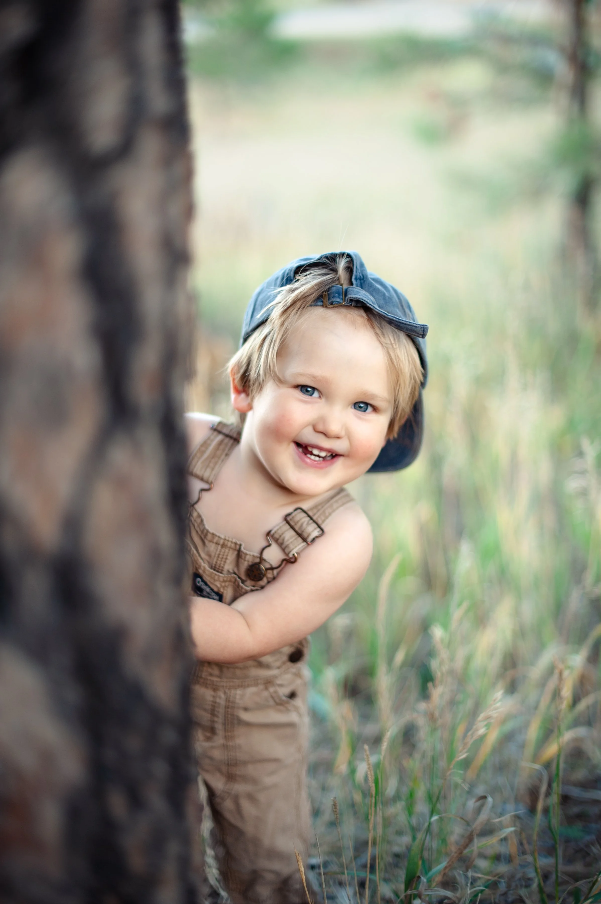 A young boy with blue eyes and blonde hair wearing a backwards blue cap and tan overalls peeks out from behind a tree in a grassy outdoor setting, smiling. Milestone childhood session, Anchorage. AK