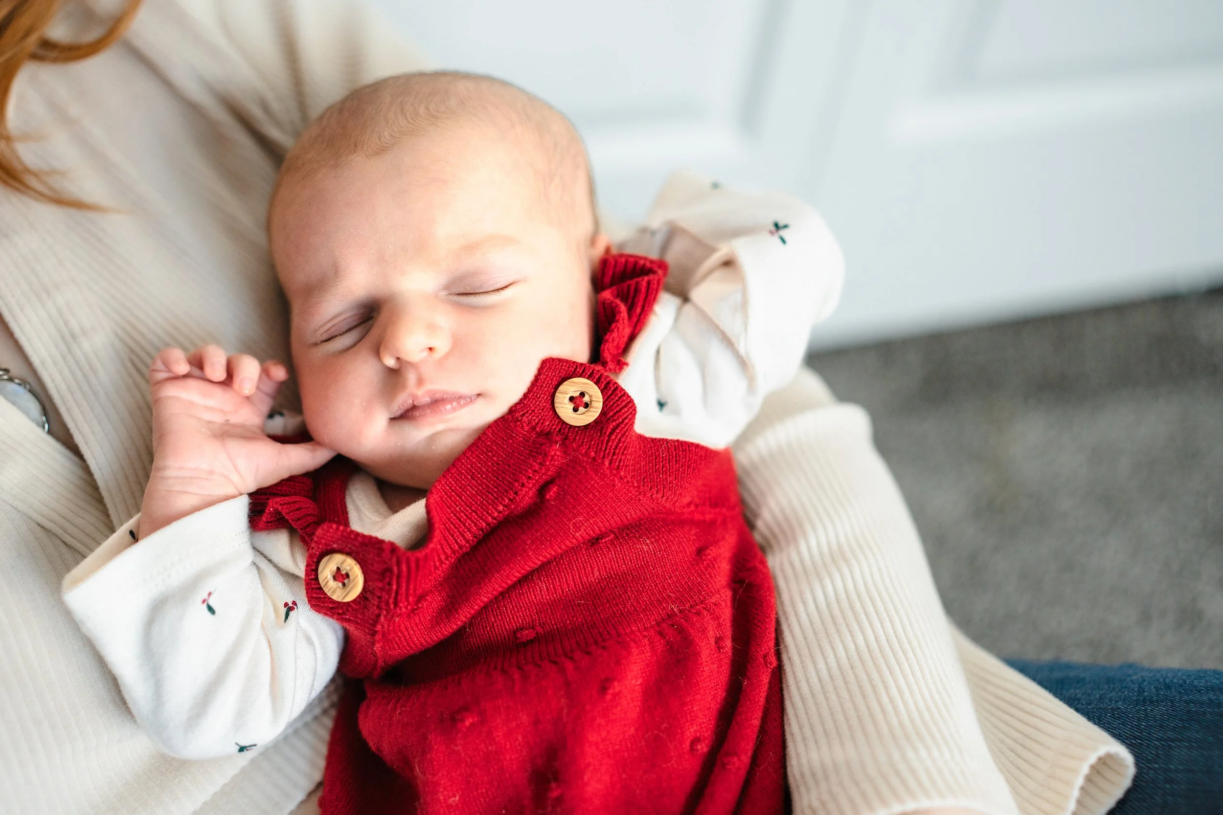 A sleeping baby dressed in a cream-colored top with small embroidered cherries, layered with a red corduroy jumper with large buttons, resting peacefully on an adult's lap. In home lifestyle newborn session Wasilla, AK.
