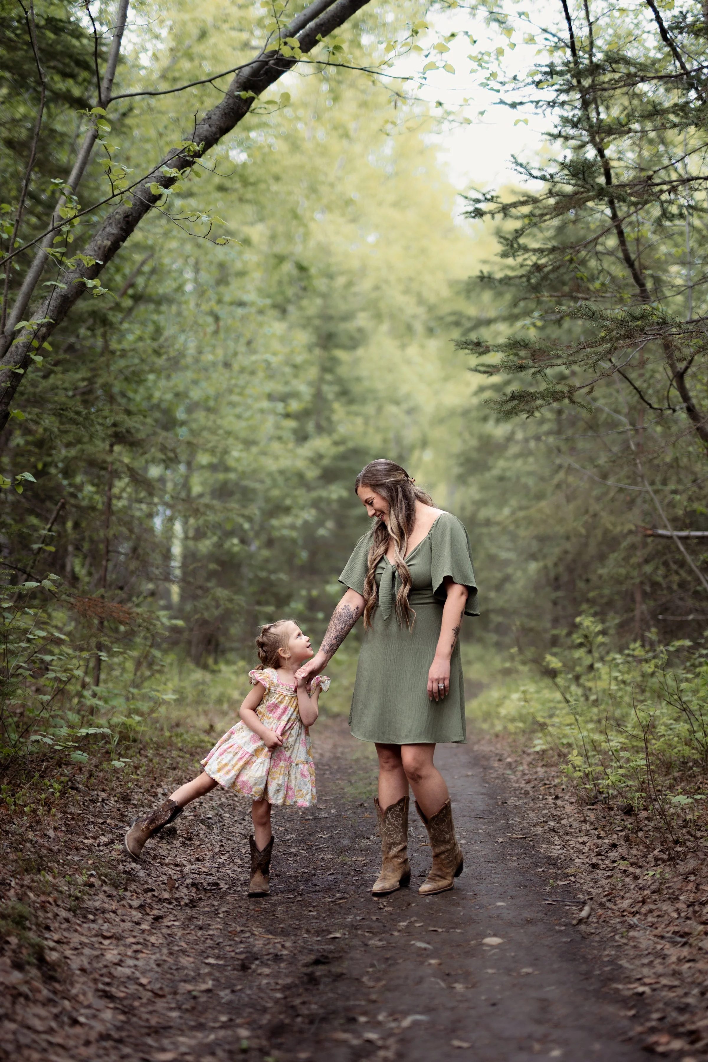 A woman and a young girl holding hands and smiling on a dirt forest trail, surrounded by green trees. Mother Daughter photoshoot in Houston, Ak.