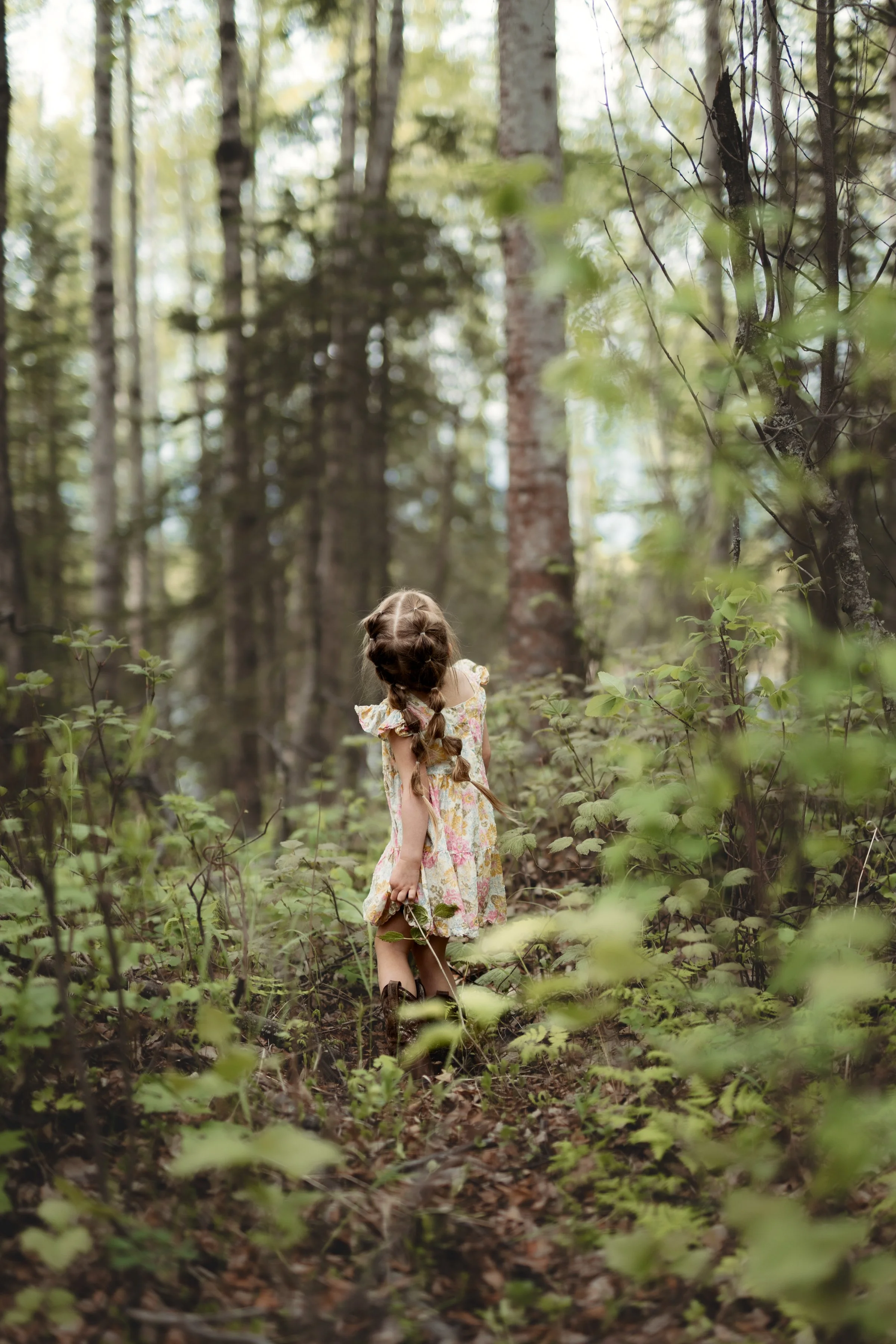 A young girl with braided hair wearing a floral dress walking through a wooded forest. Childhood photo in Girdwood, Alaska.