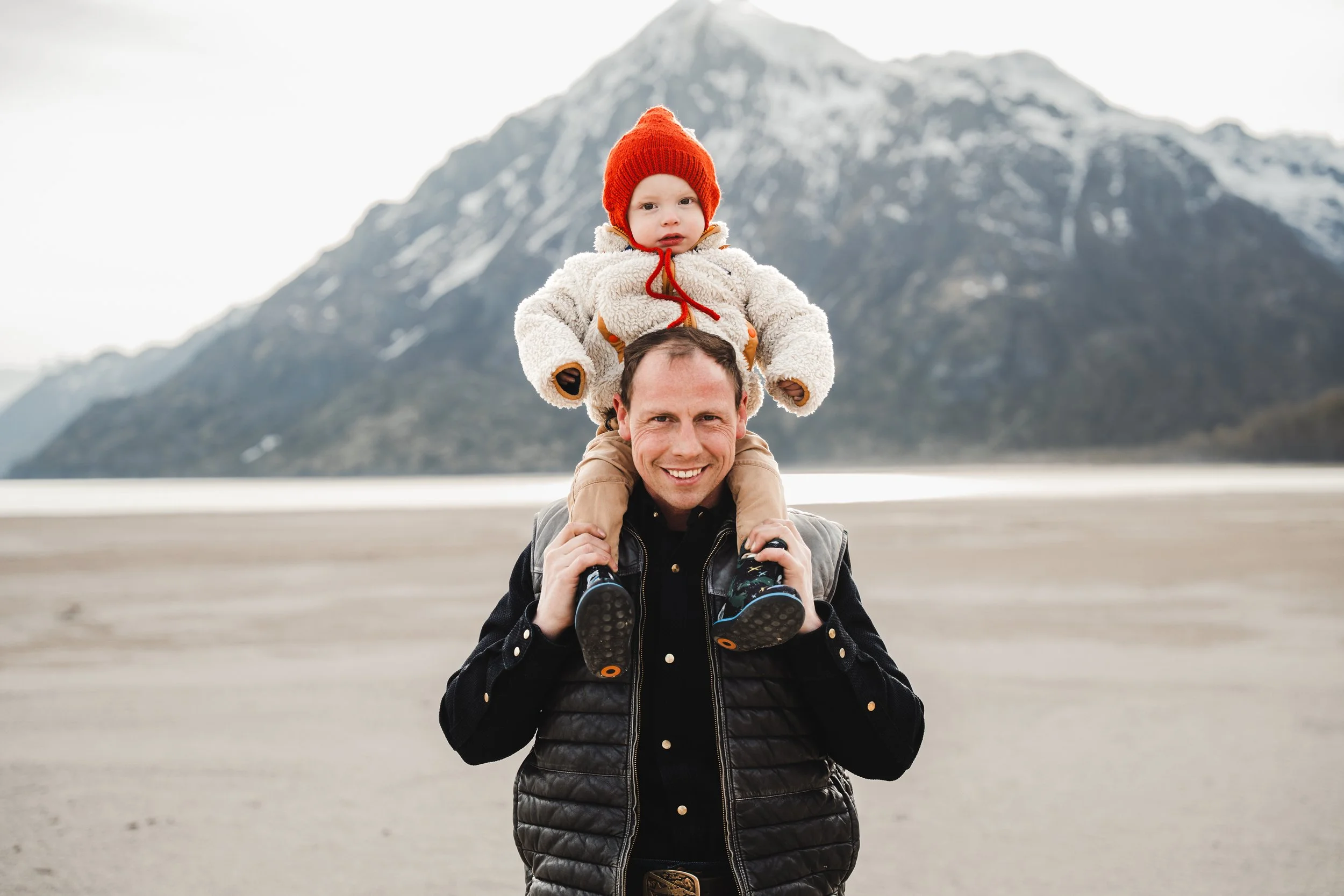 A man carrying a child on his shoulders outdoors with mountains and a body of water in the background, on a cloudy day. Big Lake, Alaska.