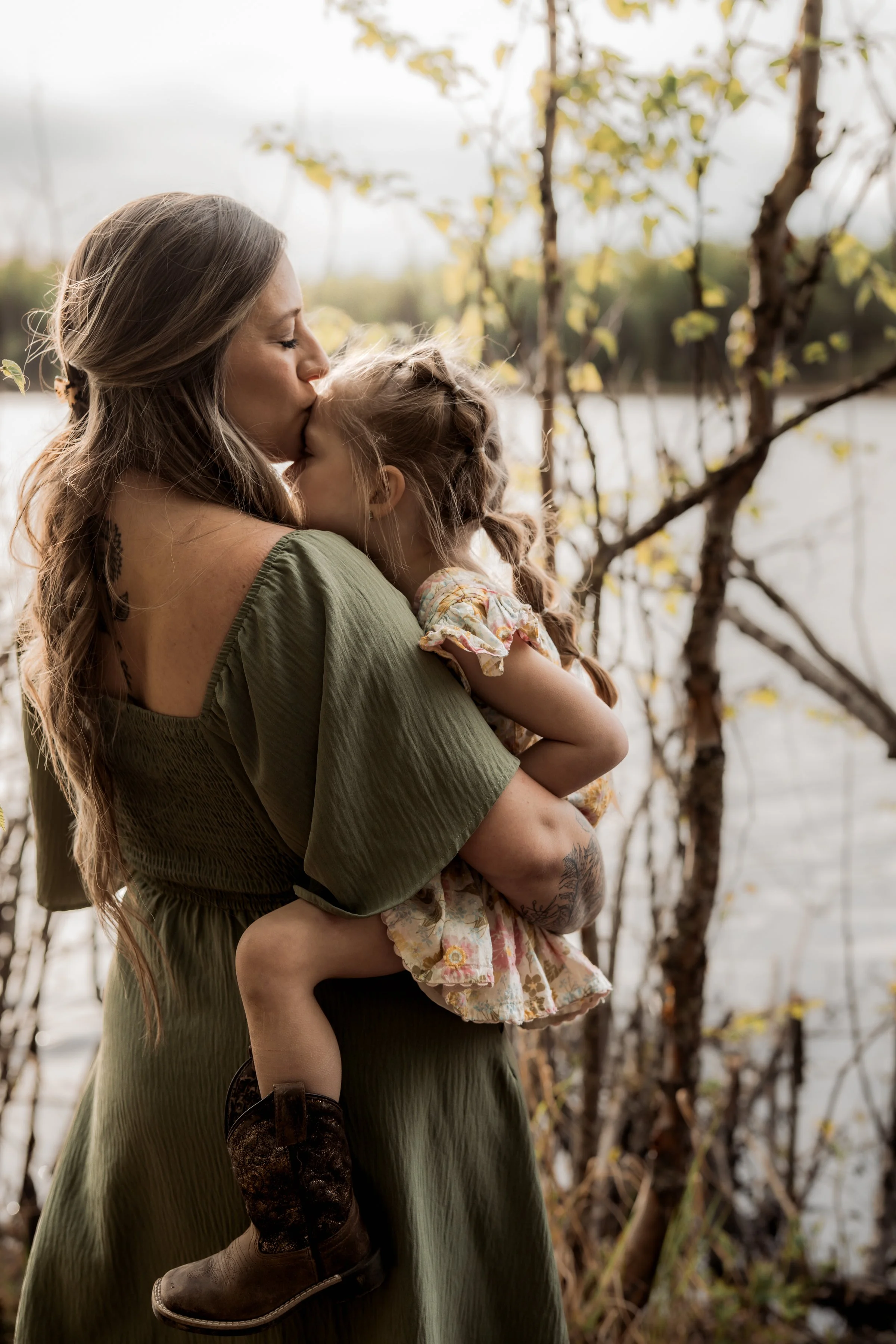 A woman with long wavy hair, wearing an olive-green dress, holding a young girl with braided hair, wearing a floral dress and cowboy boots, kissing her on the forehead outdoors near a lake with trees in the background. Reflection Lake Portraits.