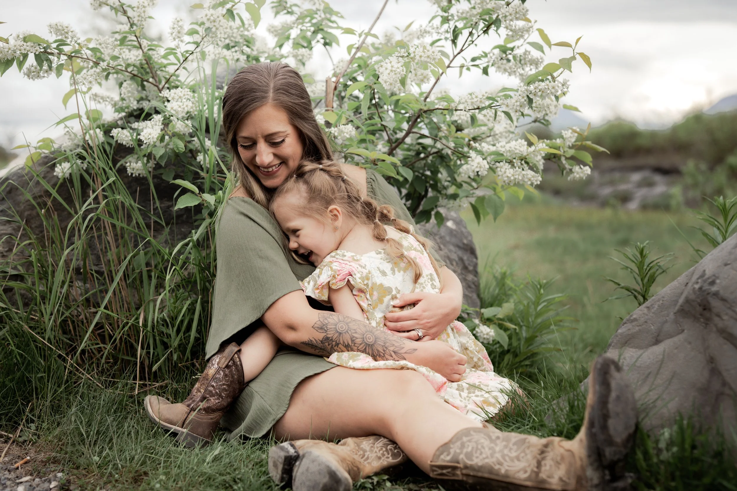 A woman with tattoos and a young girl with pigtails sharing a hug outdoors near greenery and white flowers. Motherhood photographer Palmer AK