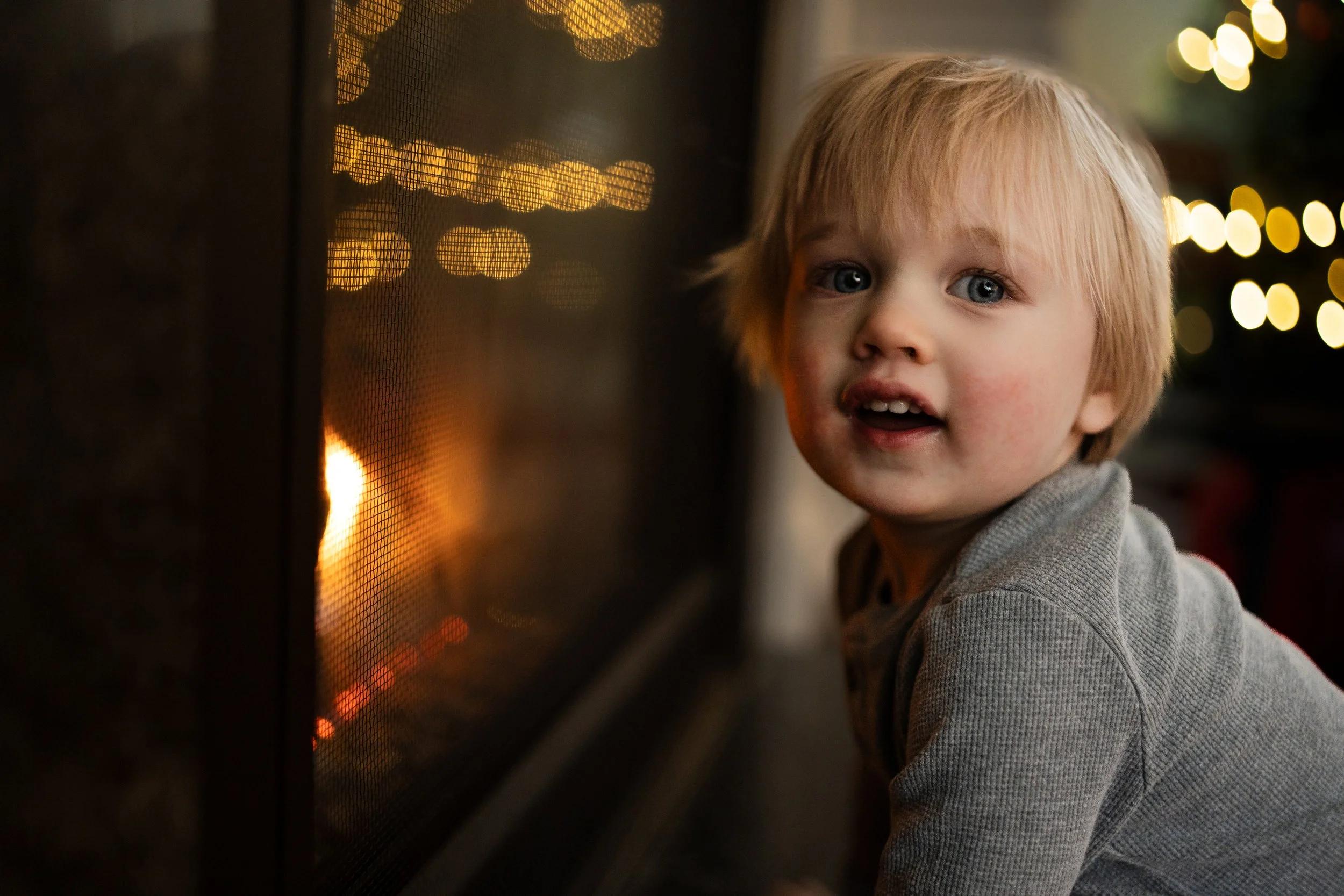 A young boy with blond hair and blue eyes watching a fire through a glass window, with holiday lights blurred in the background. In home Family lifestyle photos. Palmer, Alaska.