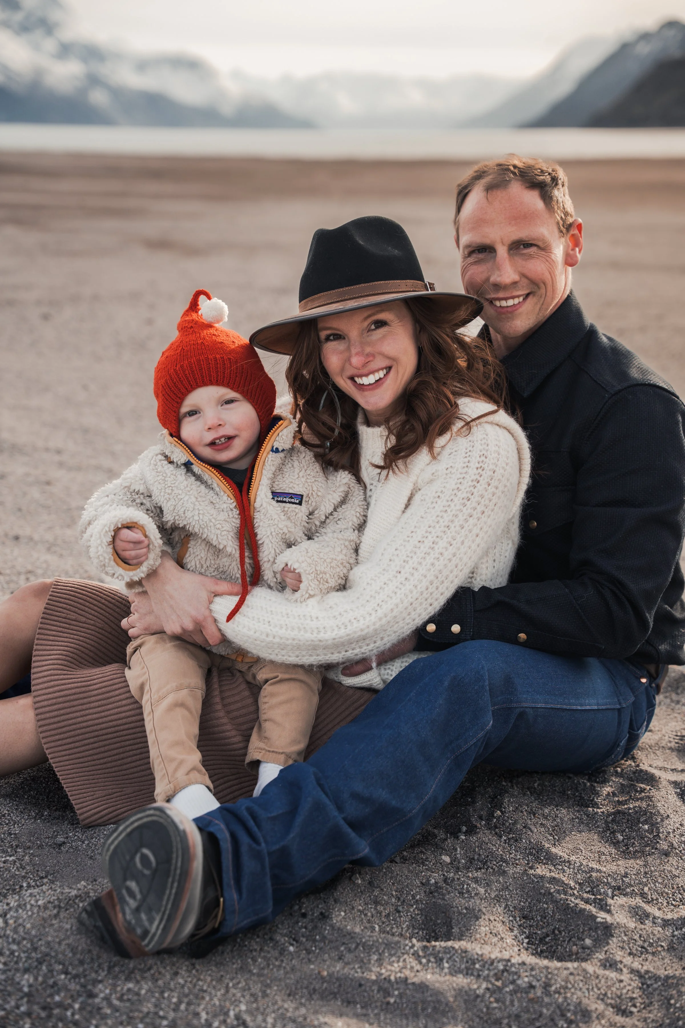 A smiling family of three sitting on the sandy beach outdoors, with mountains and a lake in the background. The woman is wearing a hat and a white sweater, the man is in a dark shirt, and the boy is in a cozy jacket and red knit hat. Posed Portraits.