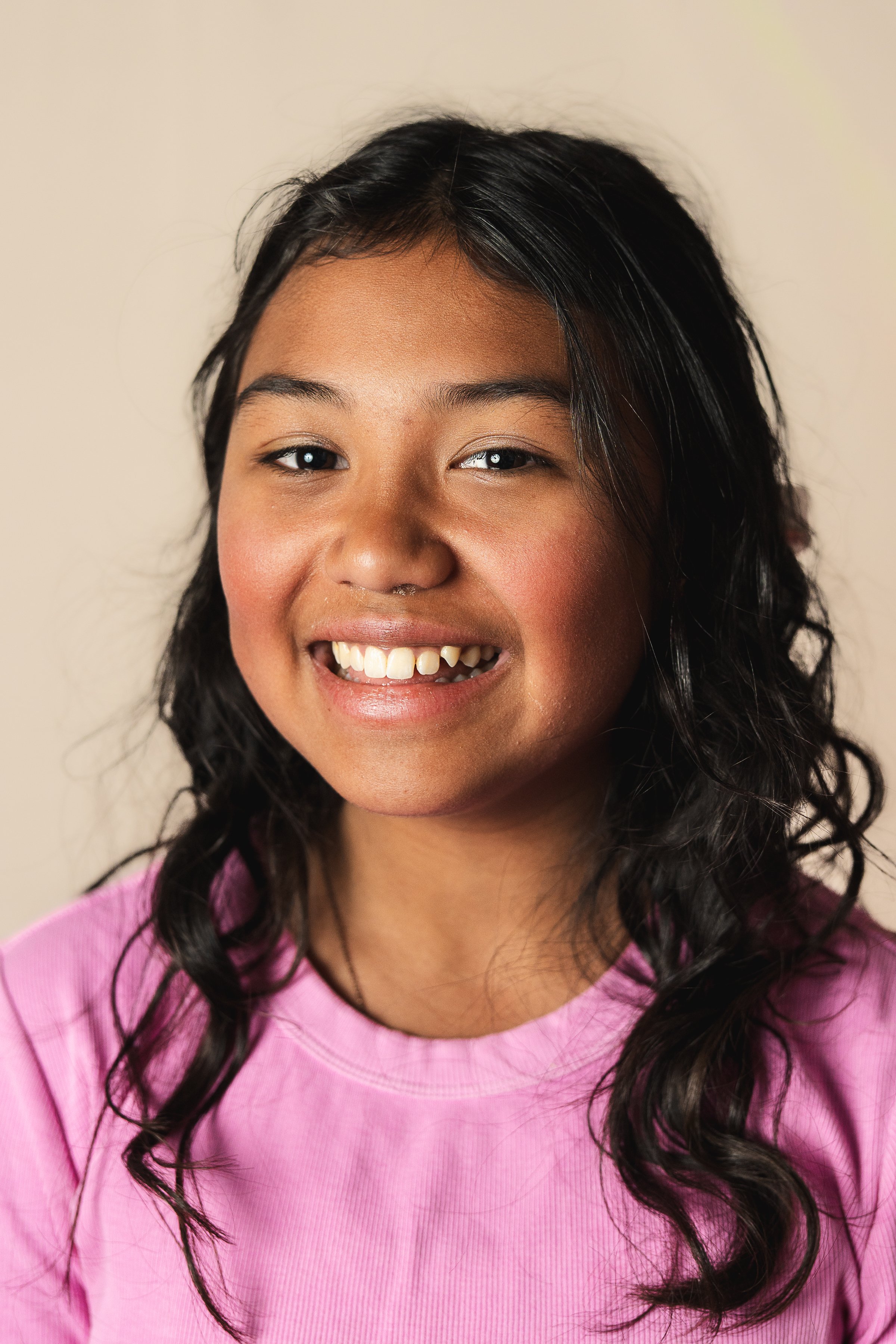 highschooler in alaska has school portrait taken on tan background. She is wearing a pink shirt and appears to be part Alaskan native. She is smiling and looking at the camera with minimal makeup and long brown curly hair.