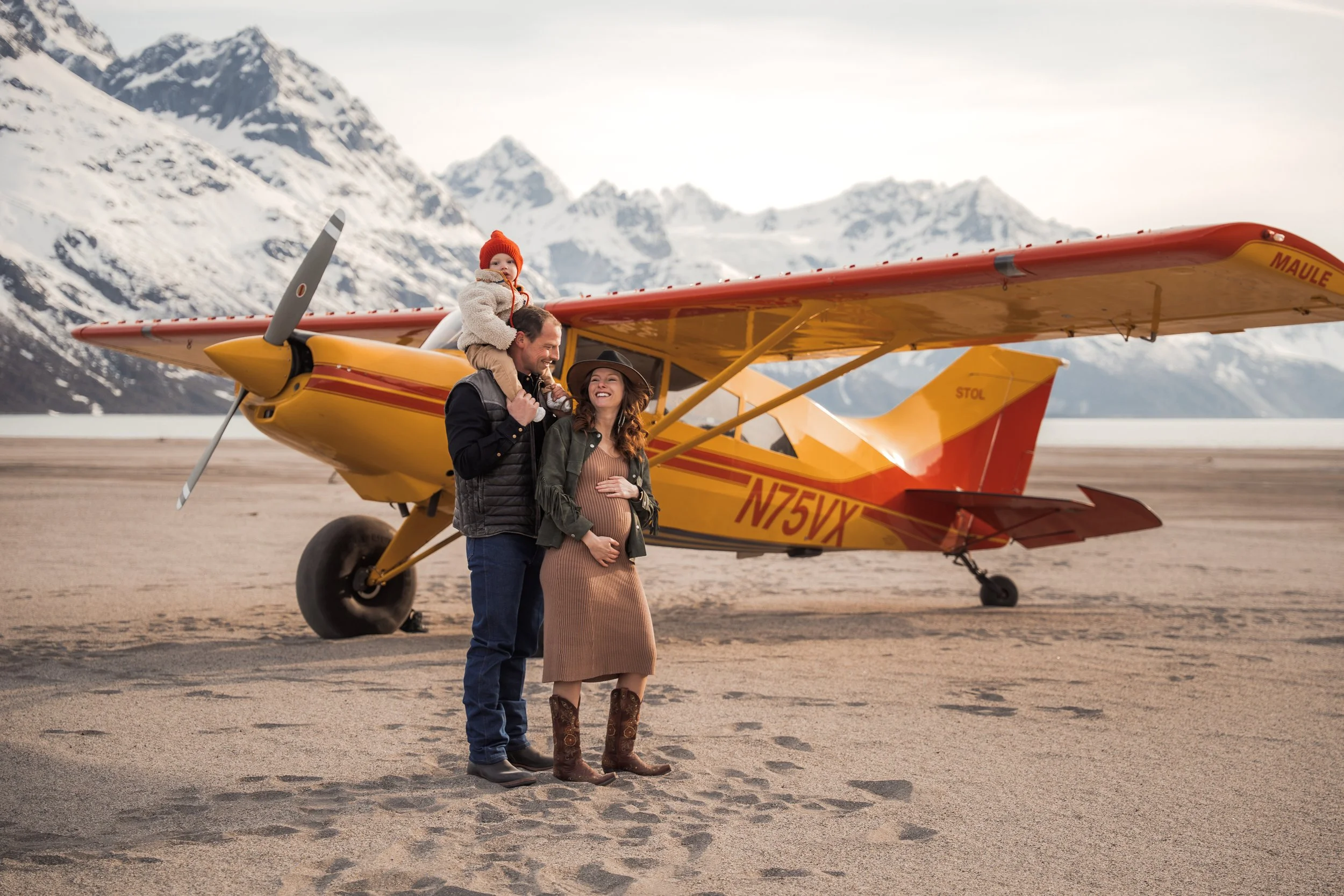 A happy family of three standing on a sandy airstrip next to a yellow and red small airplane, with snow-capped mountains in the background. and the pregnant woman is smiling and holding her belly. Anchorage Alaska Family Photos