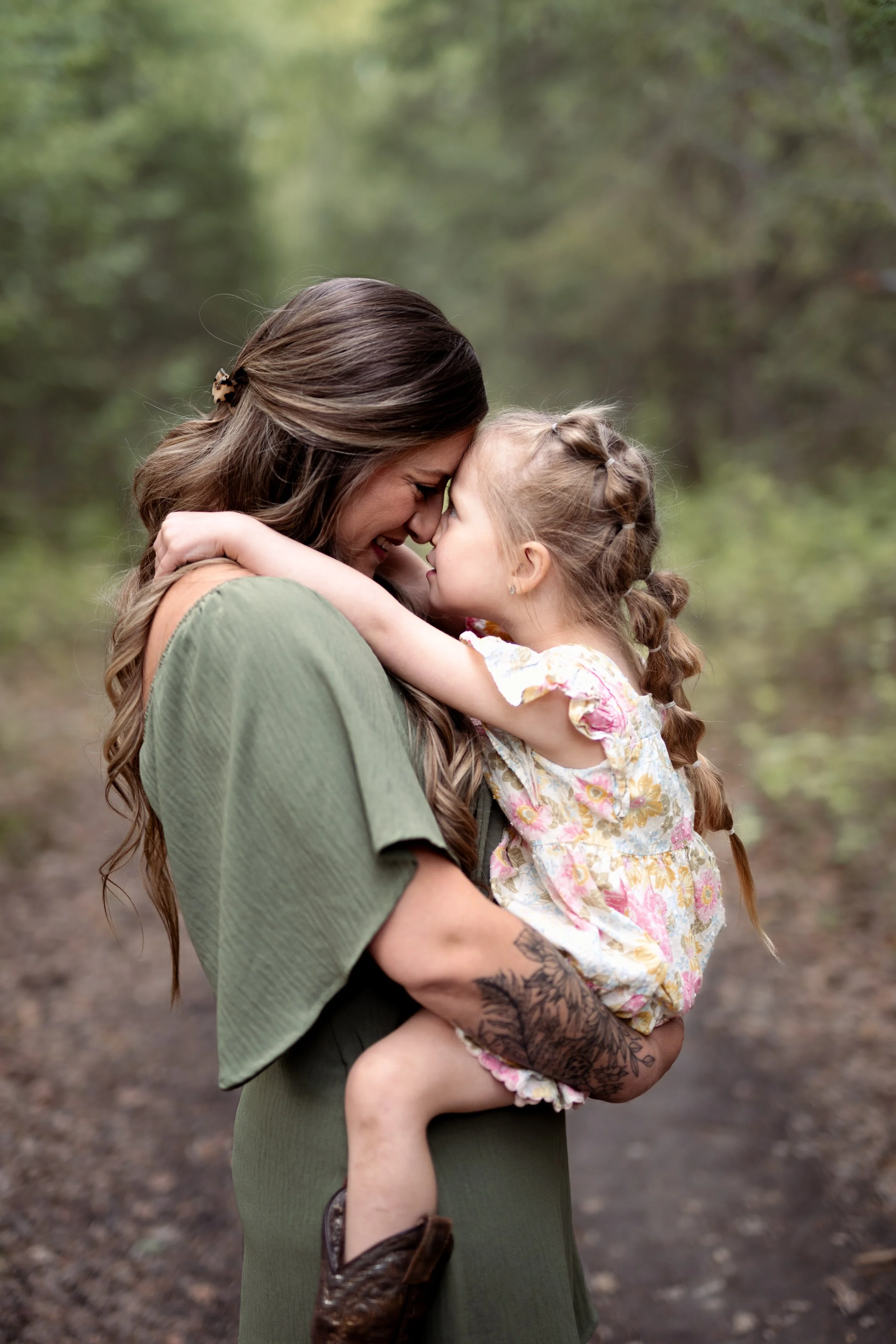 A woman with long brown hair and a tattoo on her arm is holding a young girl in a floral dress close to her face in an outdoor wooded area, both touching foreheads and smiling in Wasilla, AK.