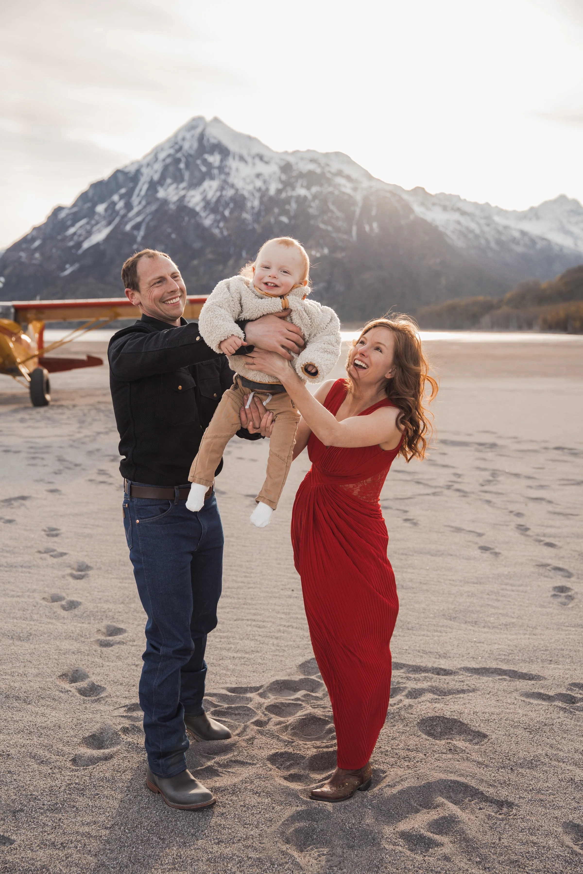 A family of three on a sandy outdoor area with mountains in the background. The man: wearing black jacket and jeans, holding the child; the woman: wearing a red dress, reaching for the child; the child: Adventurous family photos in Alaska.