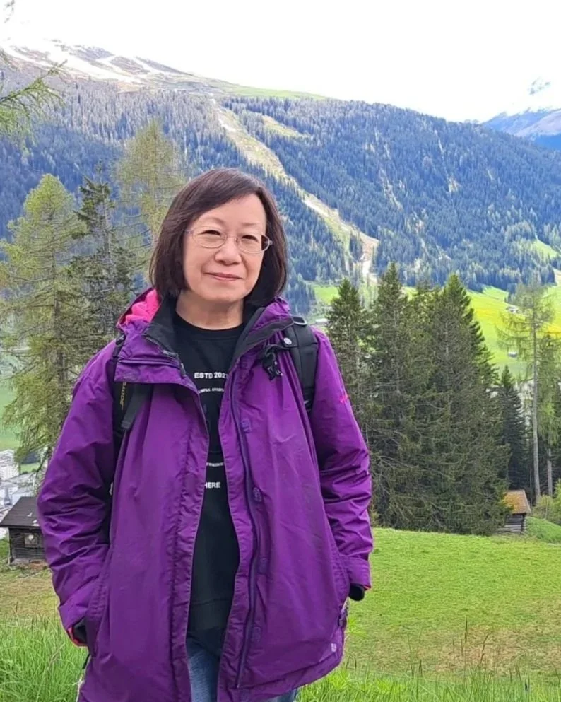 Linda To in a purple jacket standing outdoors in a grassy area with a mountainous forested landscape in the background.