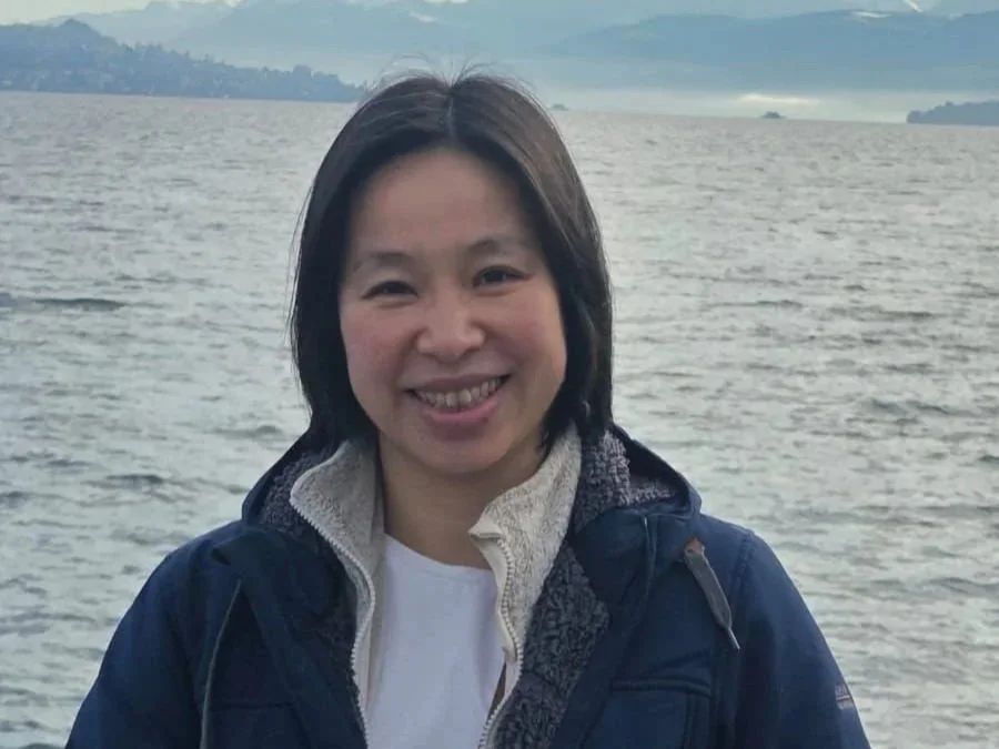 A woman smiling outdoors near a body of water with mountains in the background.