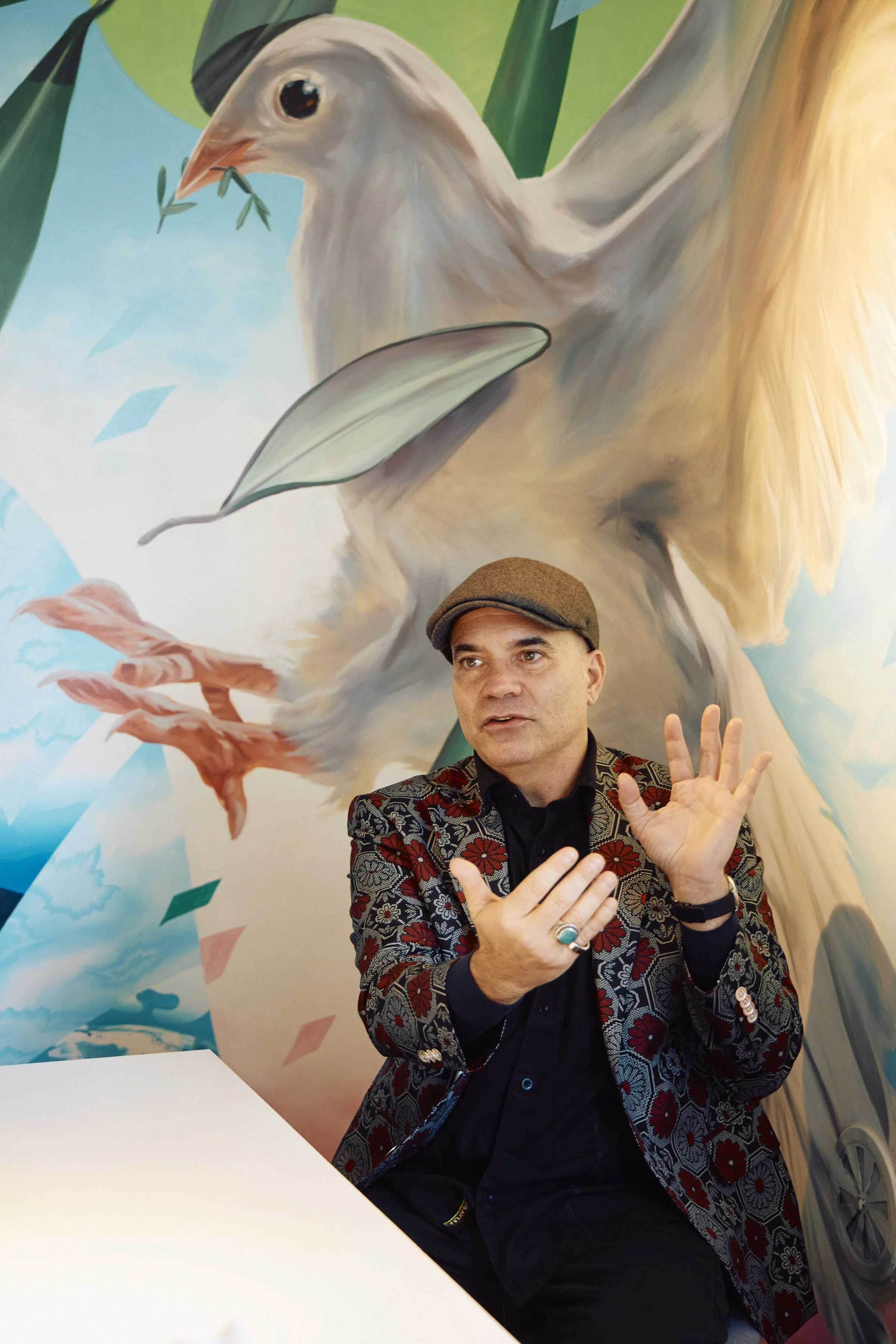 Daniel wearing a patterned blazer and a flat cap is speaking, gesturing with his hands, in front of a large mural of a bird with a leafy branch in its beak and abstract background elements.