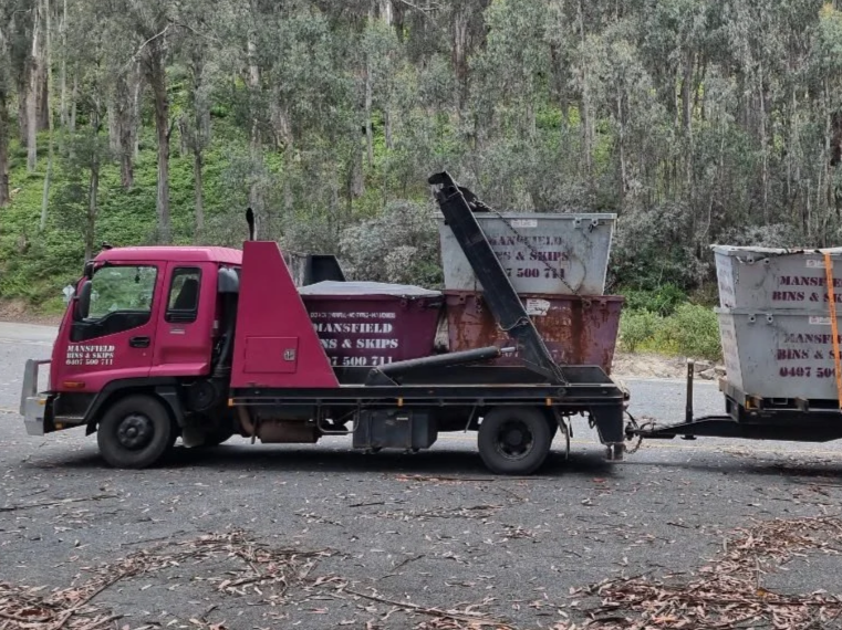A pink flatbed tow truck hauling two large industrial bins on a gravel road in a wooded area.