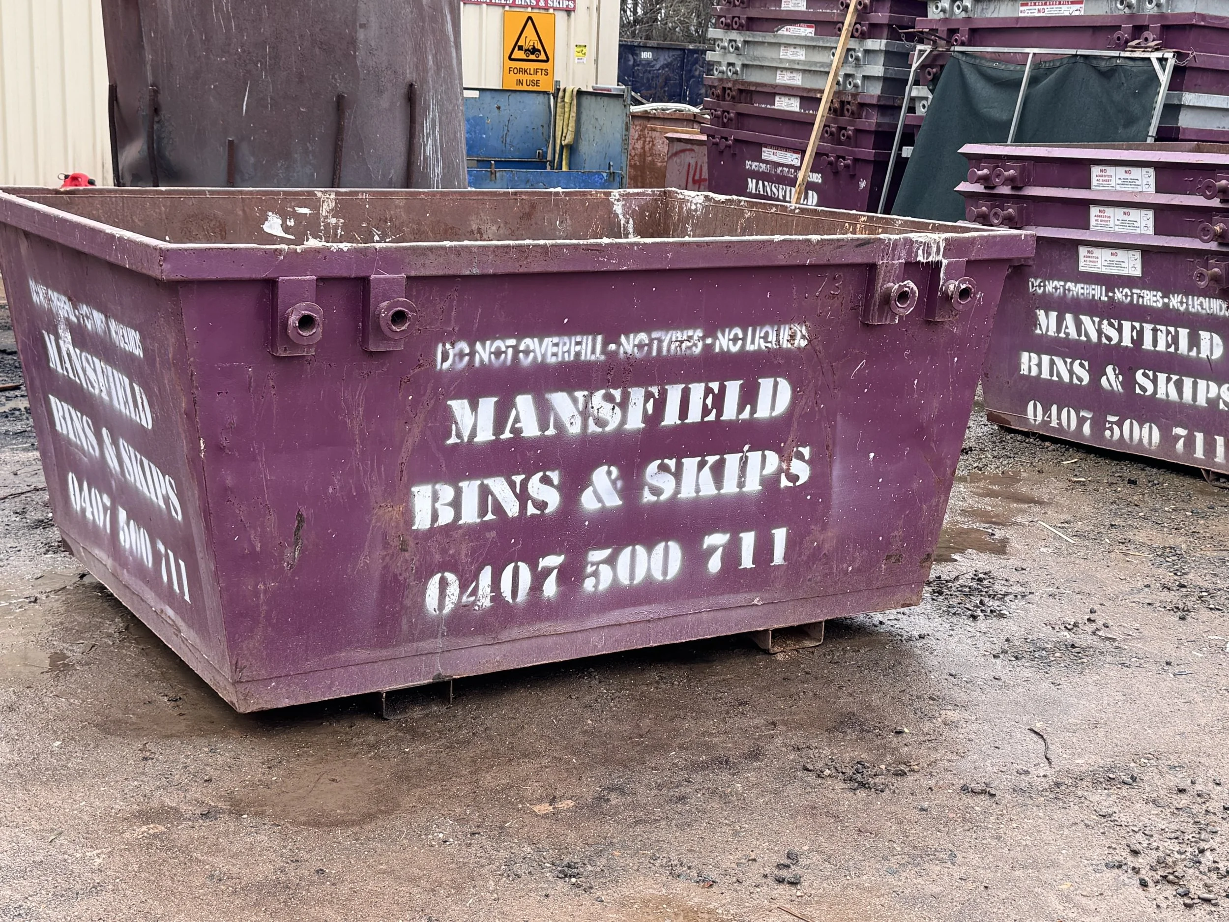 Large purple dumpster with white stencil text advertising Mansfield Bins & Skips, phone number 0407 500 711, with other dumpsters and signs in the background at a construction or industrial site.