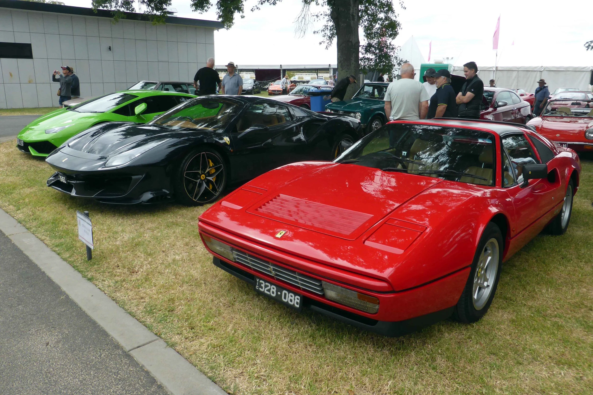 A lineup of luxury sports cars at an outdoor car show, featuring a red Ferrari 288 GTO in the foreground, a black Ferrari F430 in the middle, and a green Lamborghini Huracan in the background, with people walking and observing the vehicles.