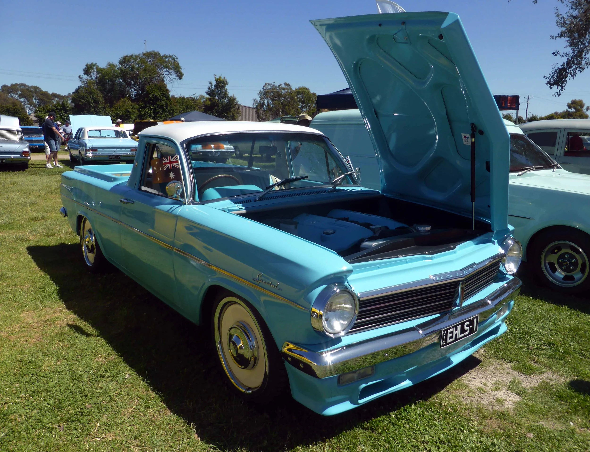 Light blue vintage Holden Special car with open hood at a classic car show on a bright sunny day, grass field, other vintage cars and people in the background.