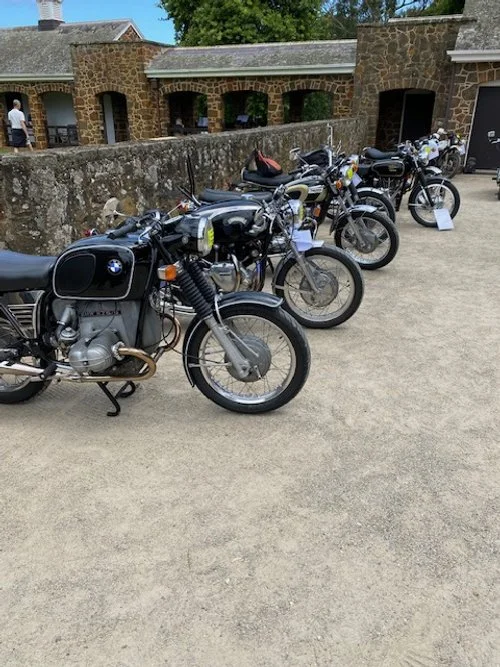 A row of vintage motorcycles parked on a concrete surface with a stone building and trees in the background.