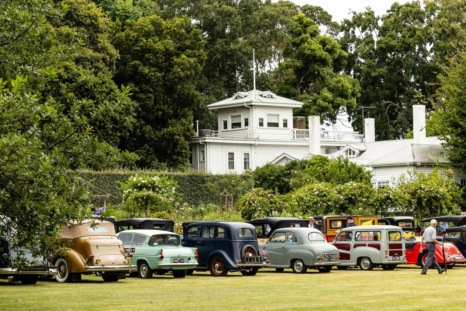 A collection of vintage cars parked on a grassy field with a historic white house and lush green trees in the background.