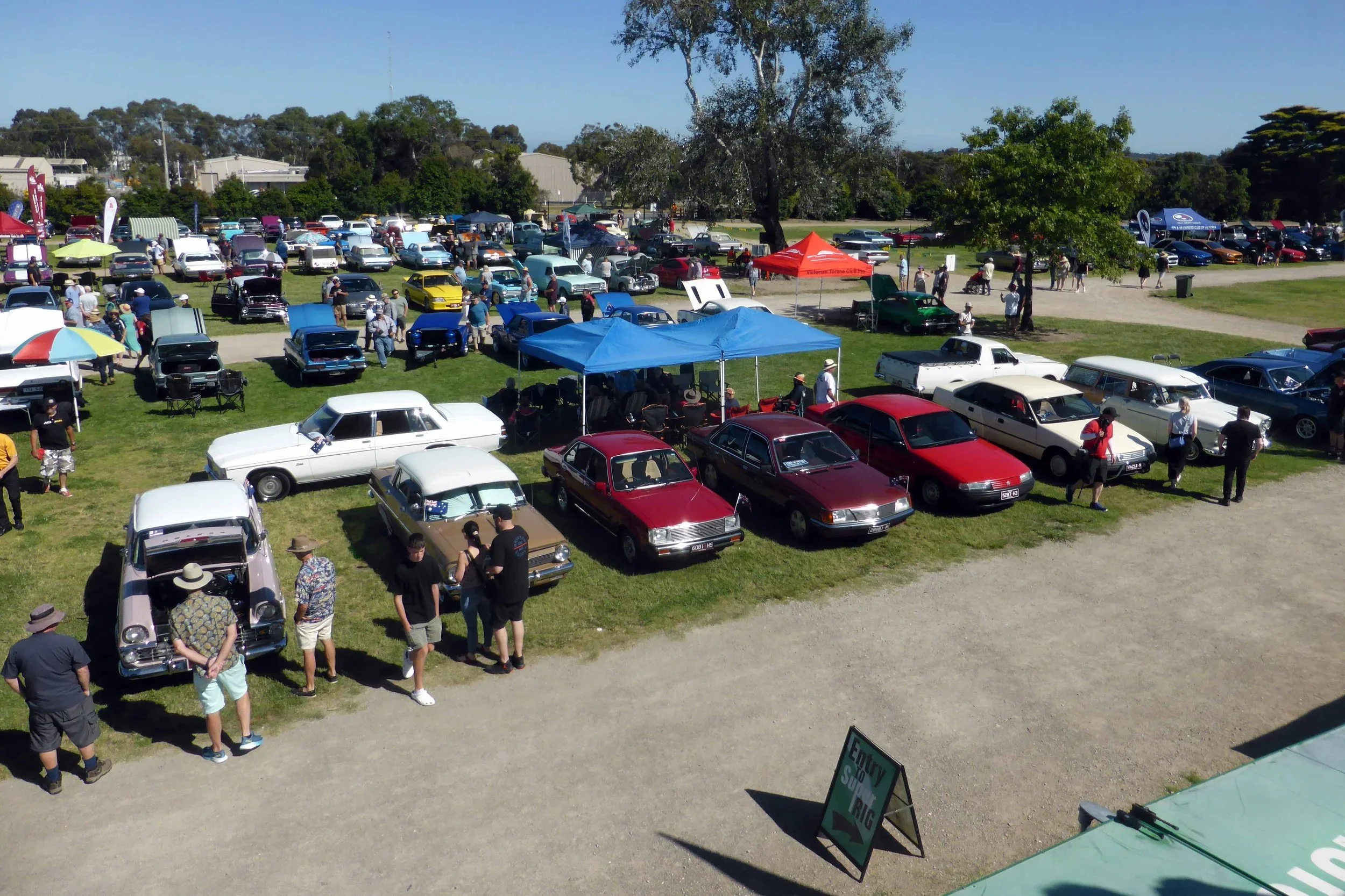 A vintage car show featuring various classic cars parked on grass and gravel, with people walking around and viewing the vehicles, and several tents and canopies set up for shade.