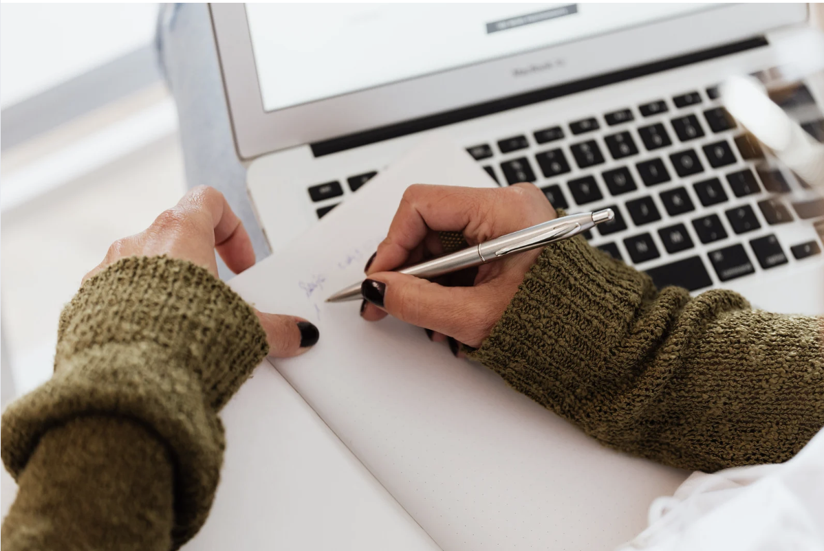 A person wearing a green sweater writing in a notebook with a pen, with a laptop nearby.