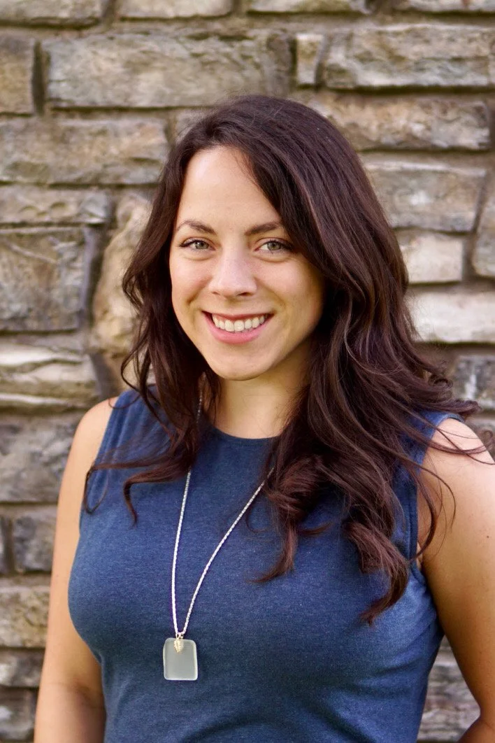 A woman with long dark brown curly hair, wearing a sleeveless blue top and a pendant necklace, stands in front of a stone wall, smiling at the camera.